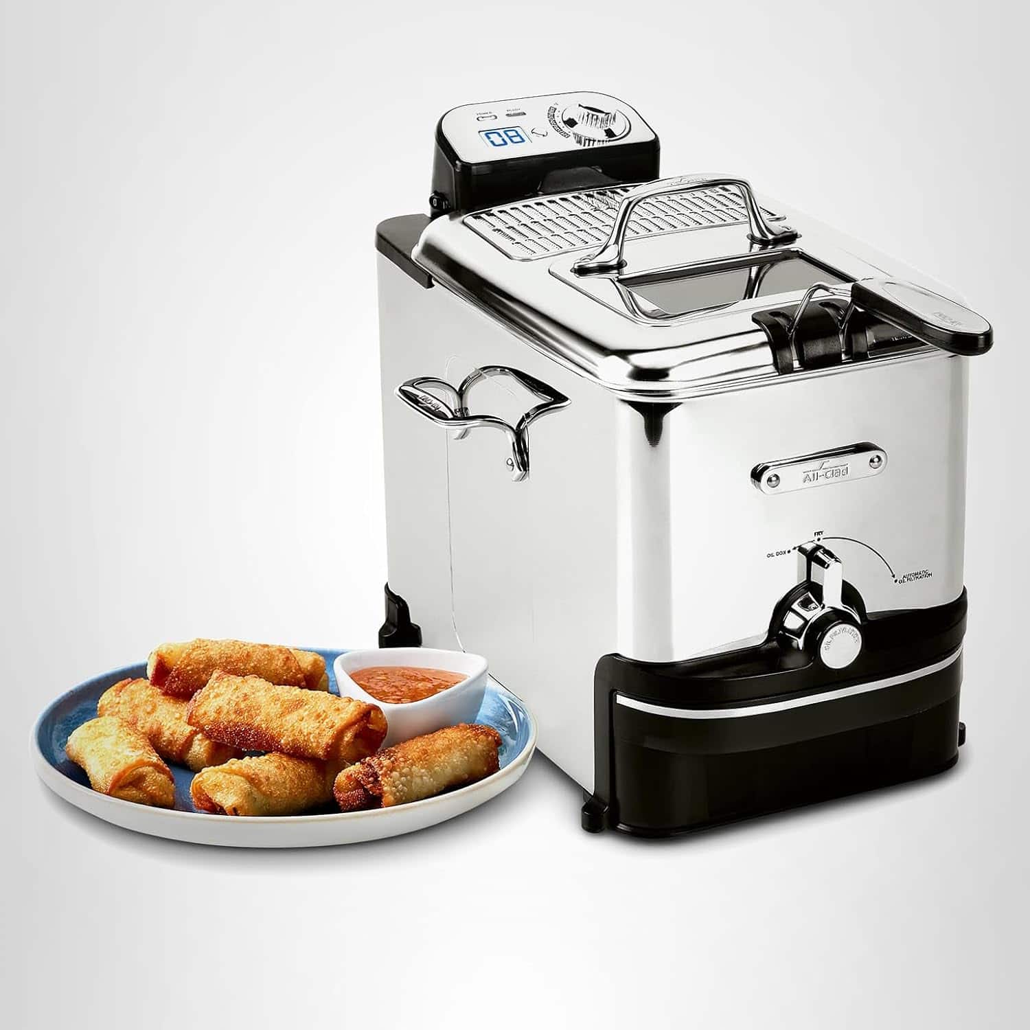 A stainless steel deep fryer is placed next to a blue plate of fried spring rolls and a small bowl of dipping sauce on a plain surface.