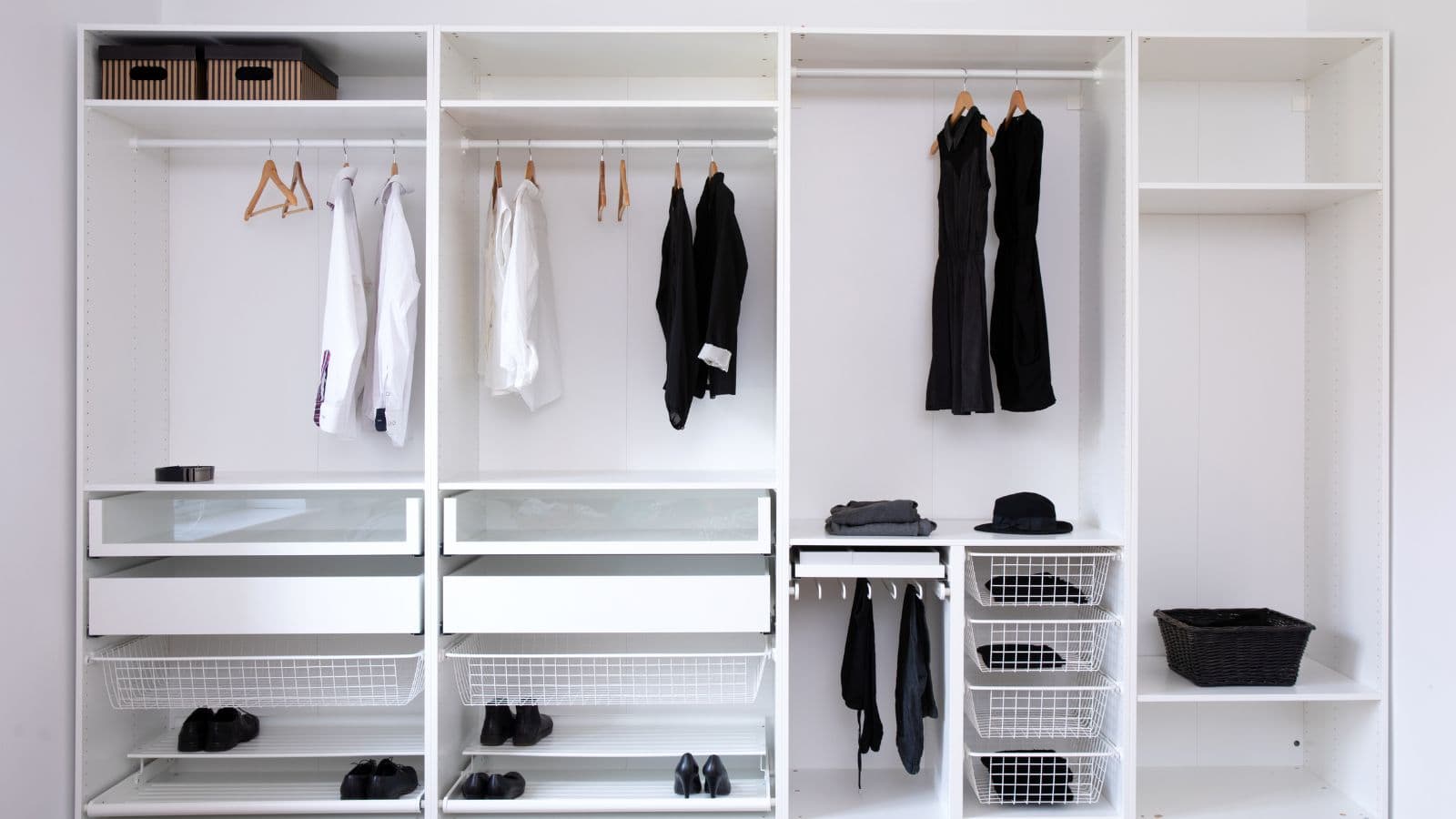 Open white closet with hanging white and black clothing, several empty hangers, shelves with shoes, baskets, and storage boxes neatly organized.
