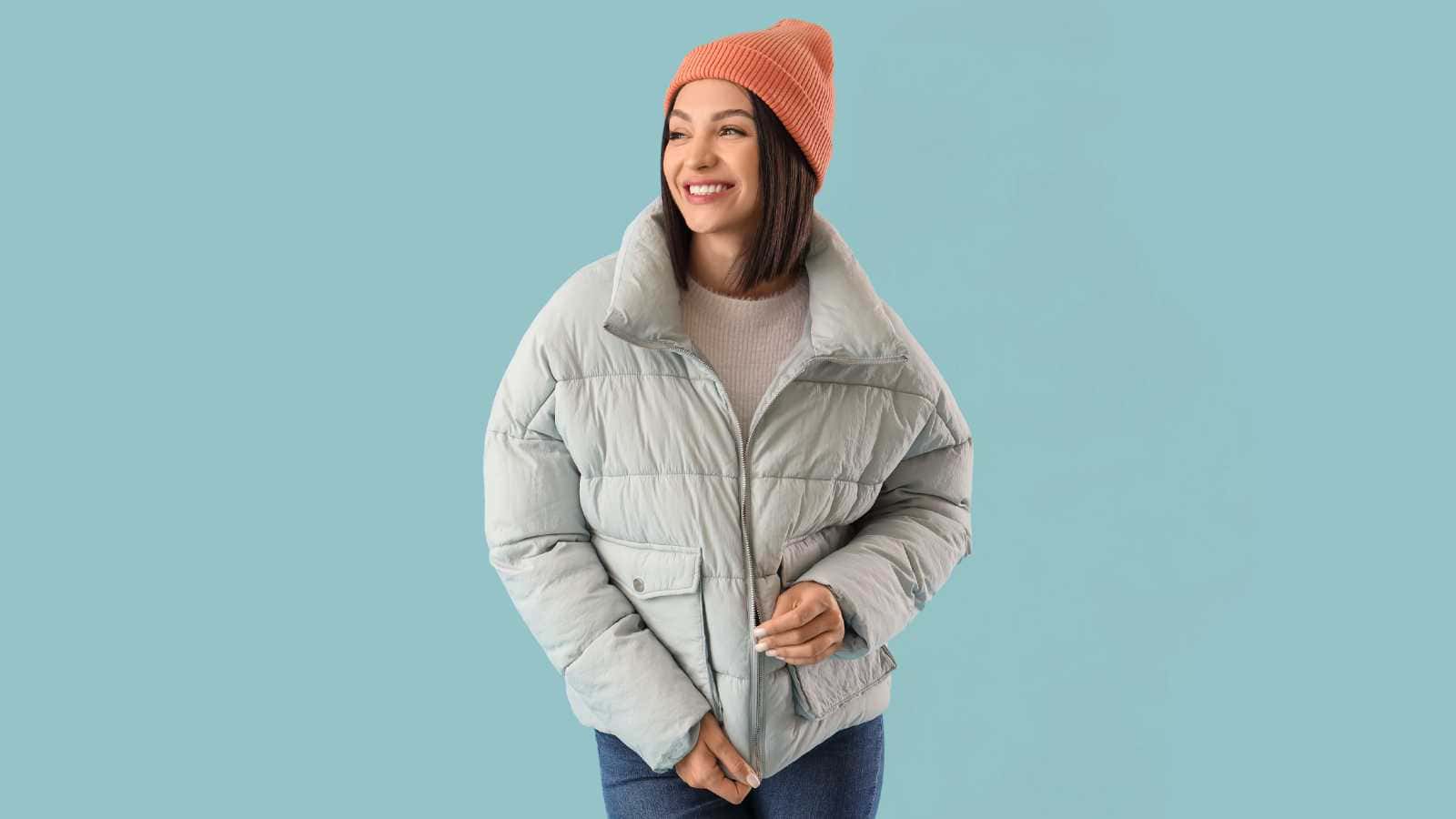 Woman in a light gray puffer jacket, orange beanie, and jeans stands against a plain light blue background, smiling and looking to the side.