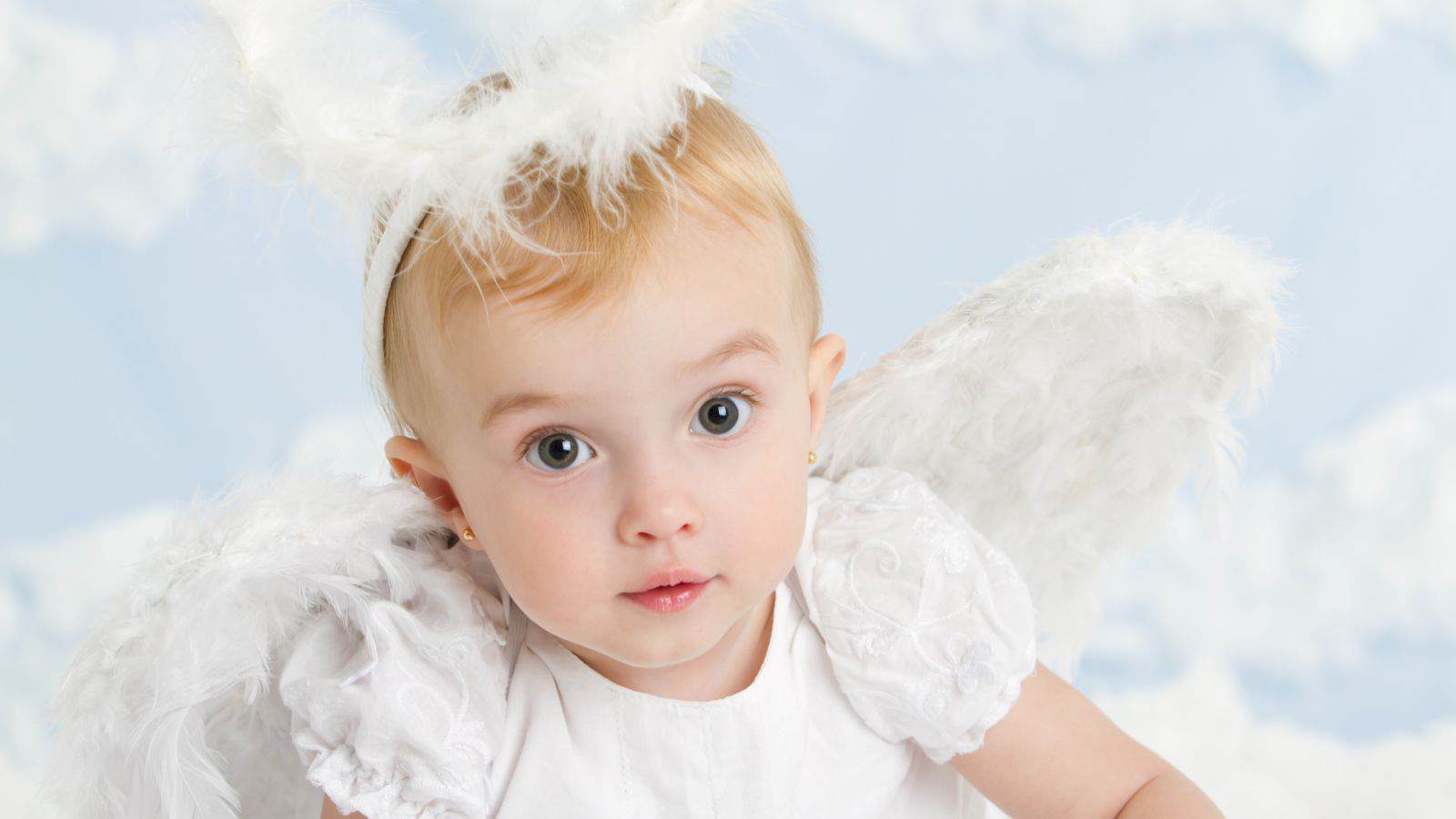 A young child dressed in a white costume with feathered angel wings and a halo headband, sitting in front of a cloud-patterned background.