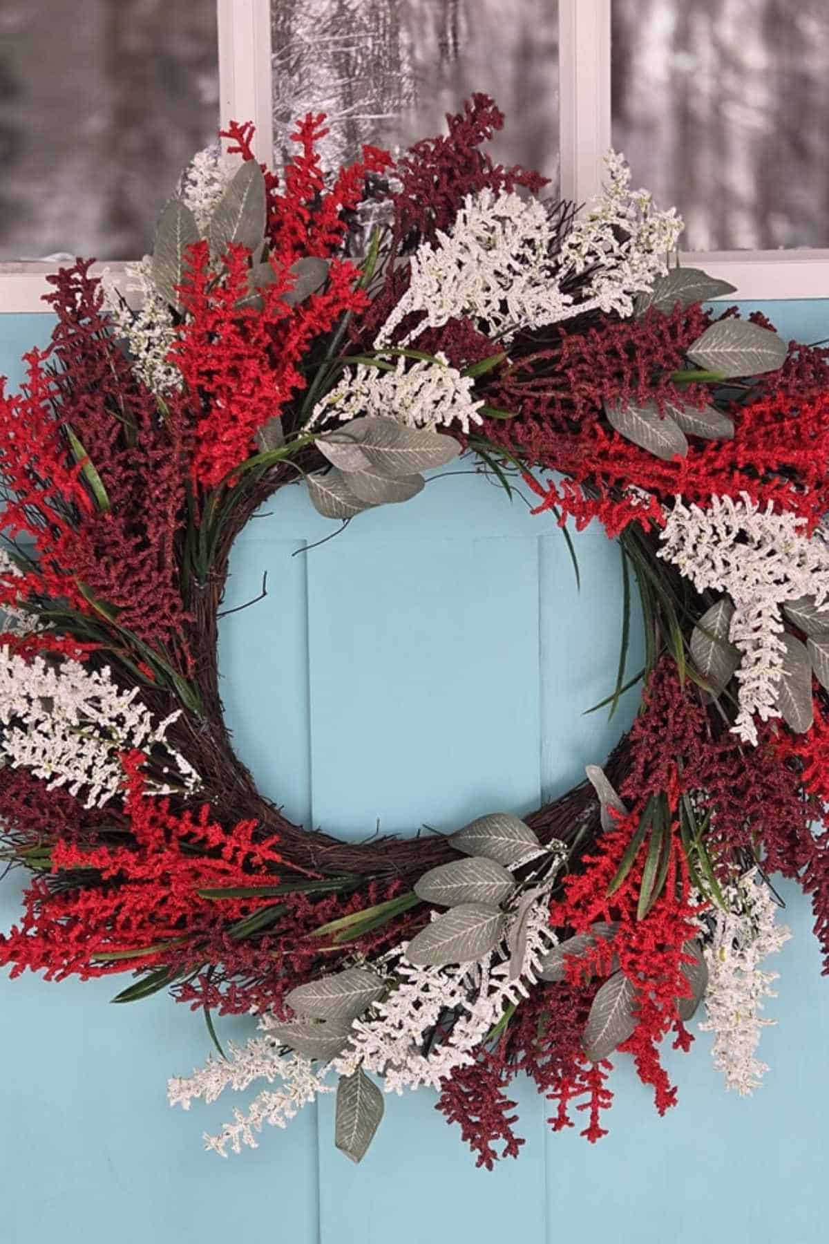 A wreath made of red and white artificial foliage with green leaves, hanging on a light blue door.