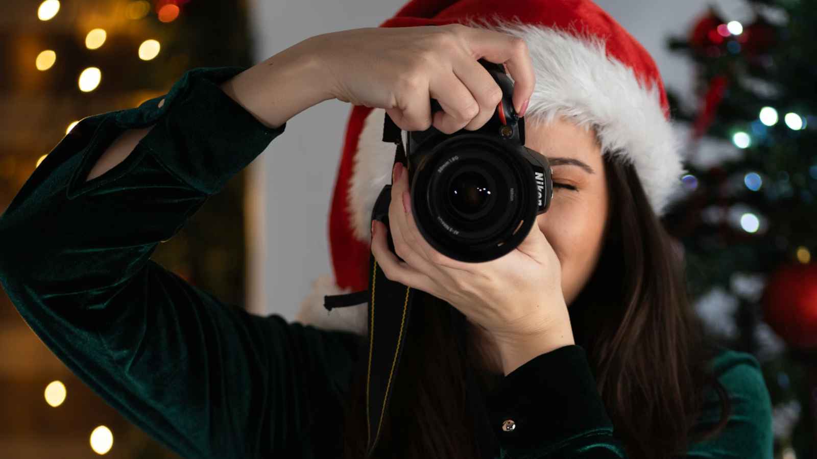 Person wearing a Santa hat and green top holds a camera, taking a photo indoors with holiday lights and a decorated Christmas tree in the background.