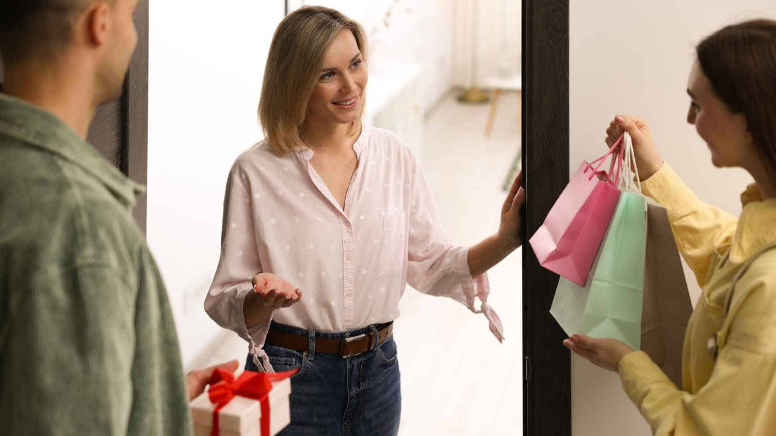 A smiling woman stands at an open door talking to two people who are holding gift bags and a wrapped present.