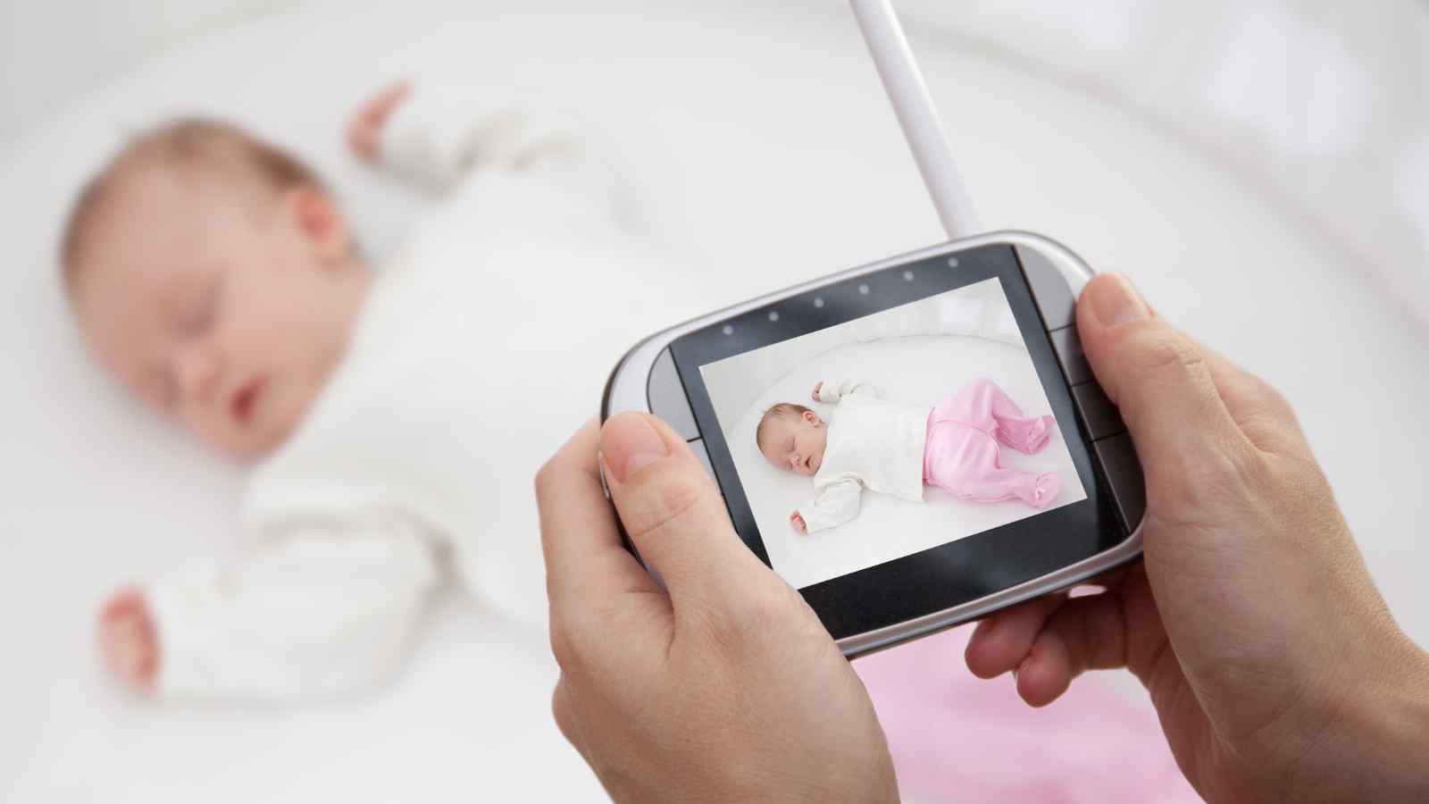 A person holds a baby monitor showing a live video feed of a sleeping baby wearing a white top and pink pants in a crib.