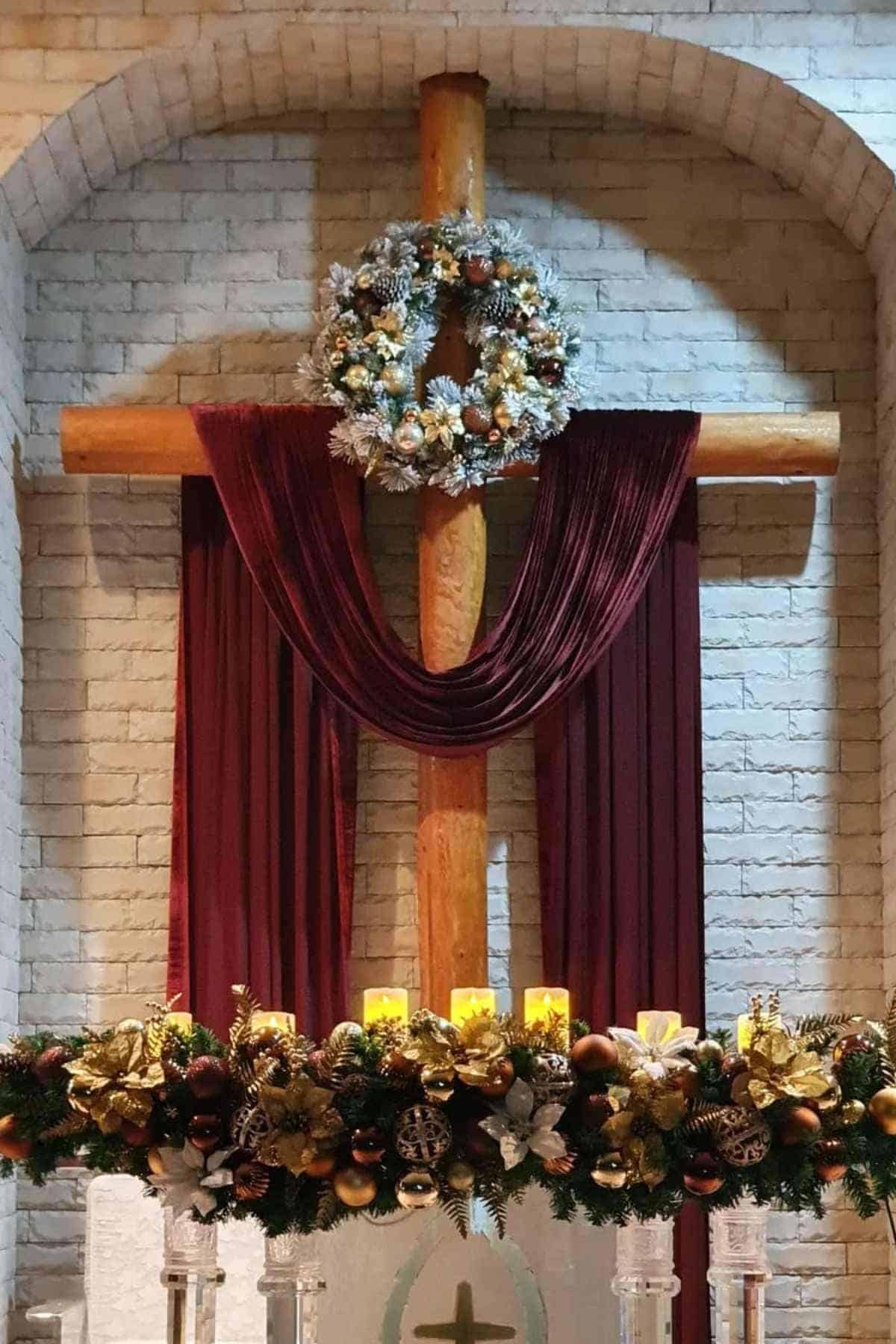 A wooden cross draped with burgundy fabric is adorned with a white Christmas wreath; below is an arrangement of gold ornaments and candles.