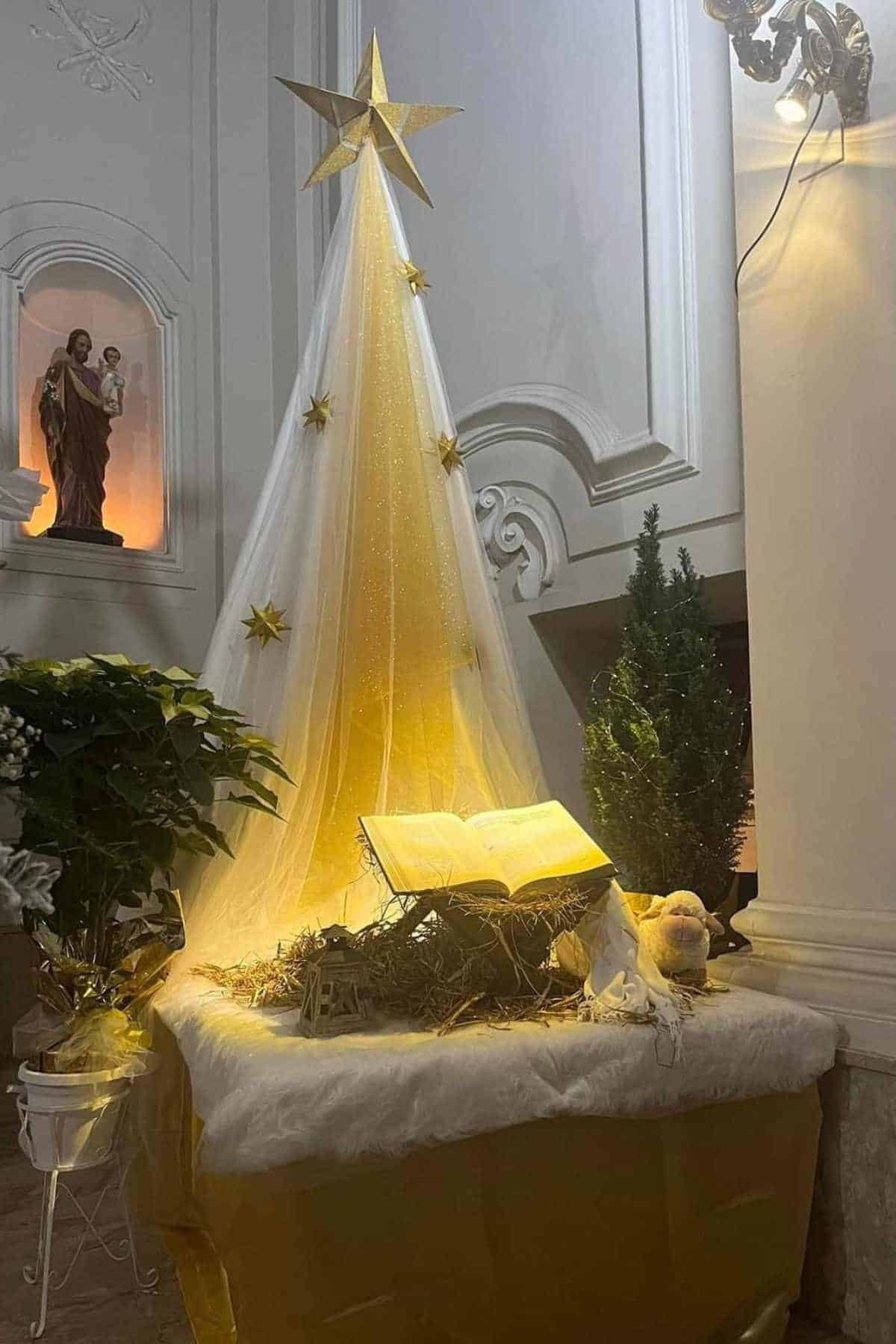 A decorative nativity display features an open book, a small tree, a plush sheep, and a white-and-gold fabric Christmas tree topped with a star, set in a church interior.