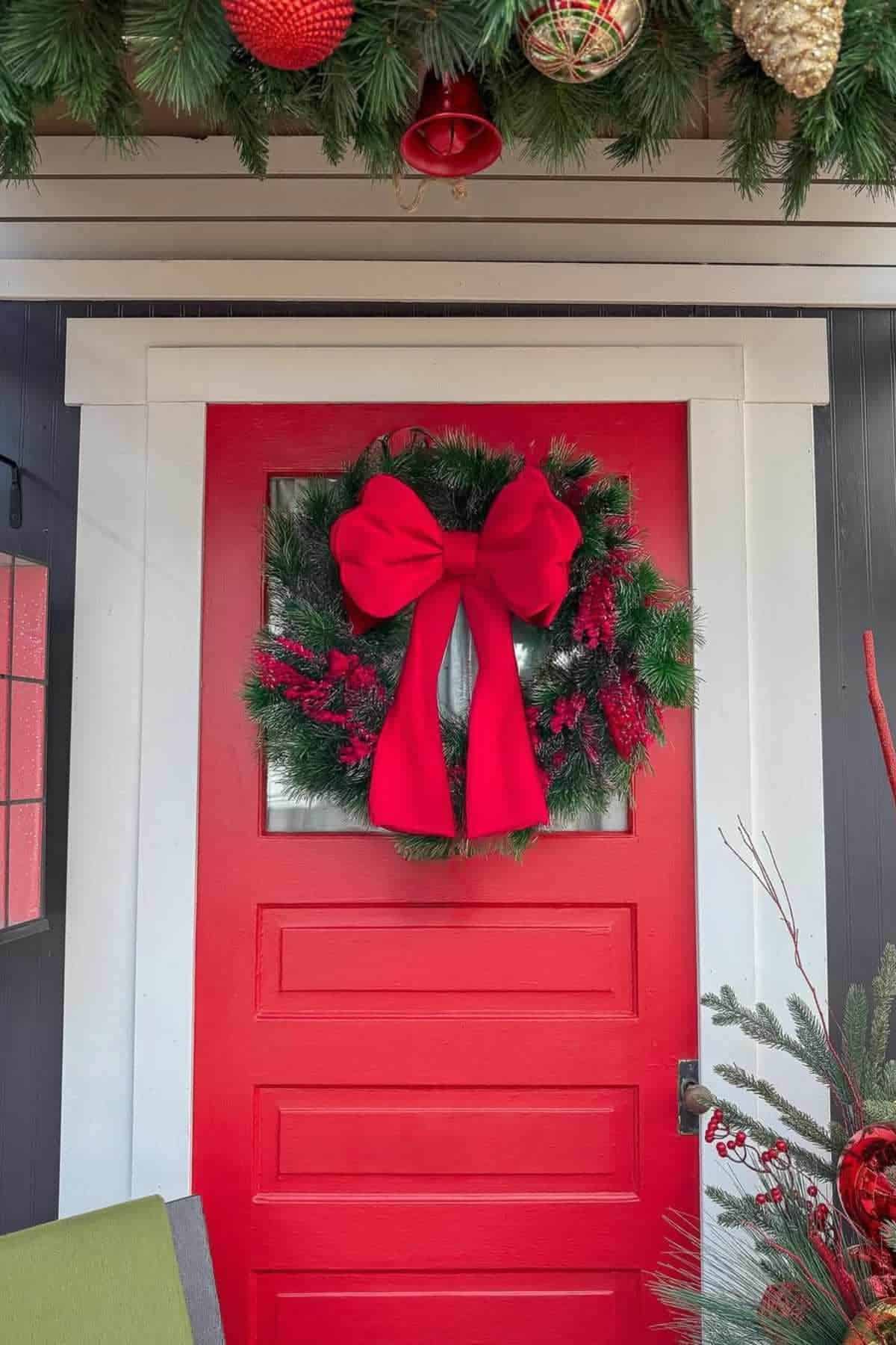 A bright red door decorated with a green wreath, red berries, and a large red bow. Pine garland and ornaments hang above the door.