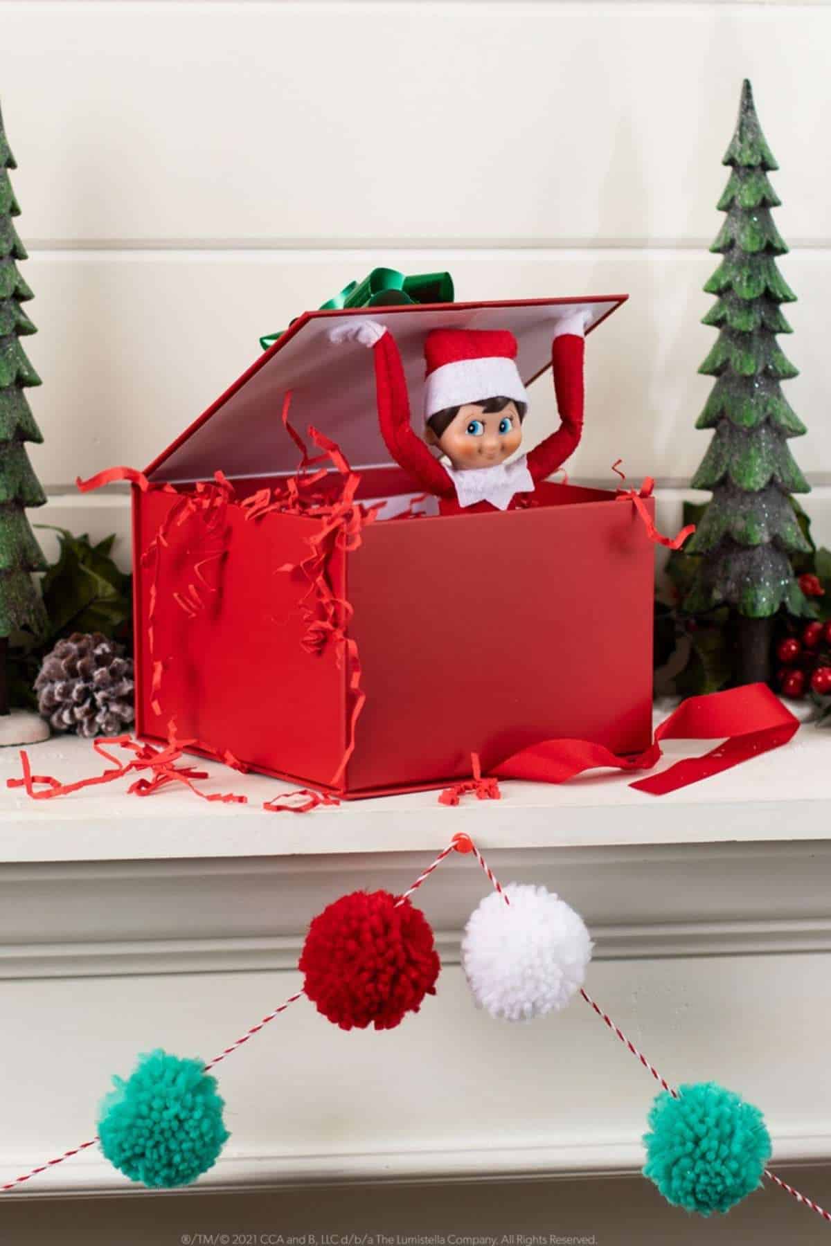 A toy elf in a red box with the lid open, surrounded by artificial trees, pine cones, and festive decorations on a mantel.
