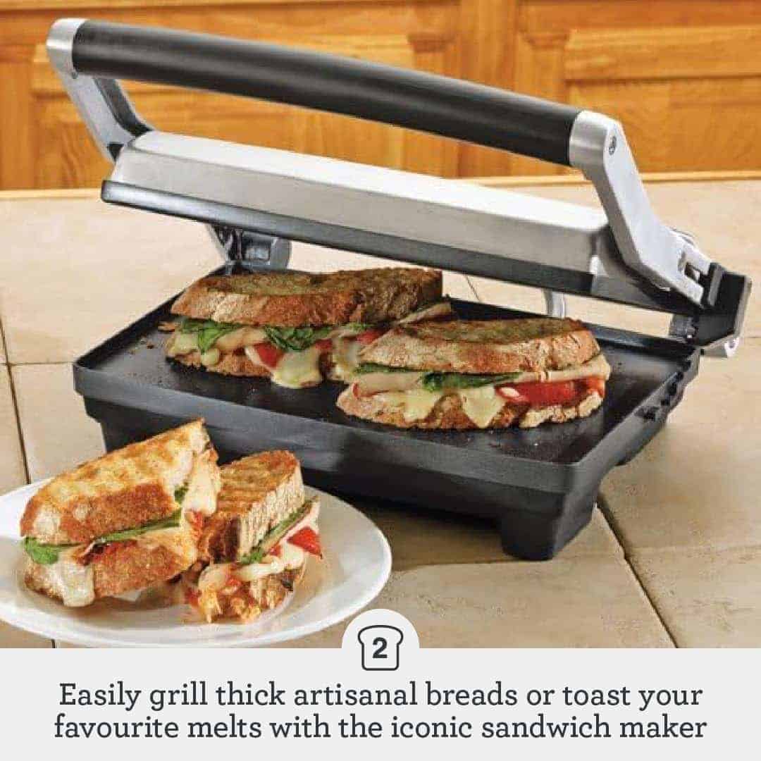 A sandwich press grilling three sandwiches with melted cheese and vegetables; two finished sandwich halves sit on the counter beside the press.