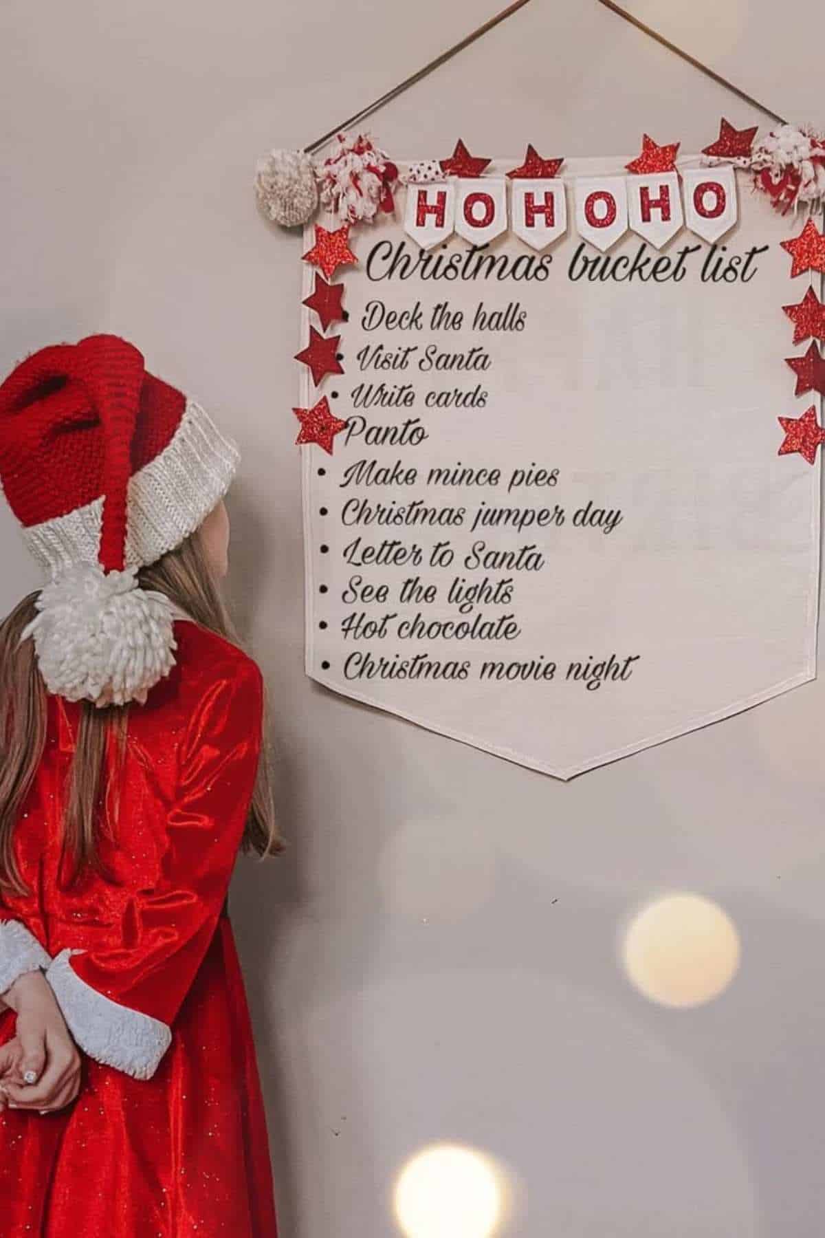A child in a red dress and Santa hat looks at a Christmas bucket list banner hanging on a wall, listing holiday activities.