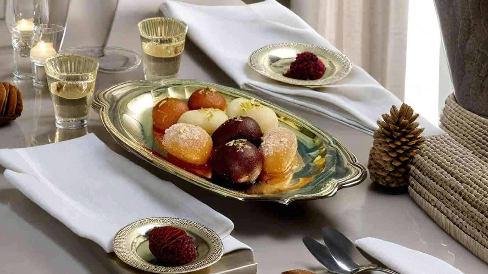 A silver tray with assorted candied fruit and two matching plates, each with a single candied fruit. Folded white napkins, glasses of clear liquid, and pinecones are also on the table.