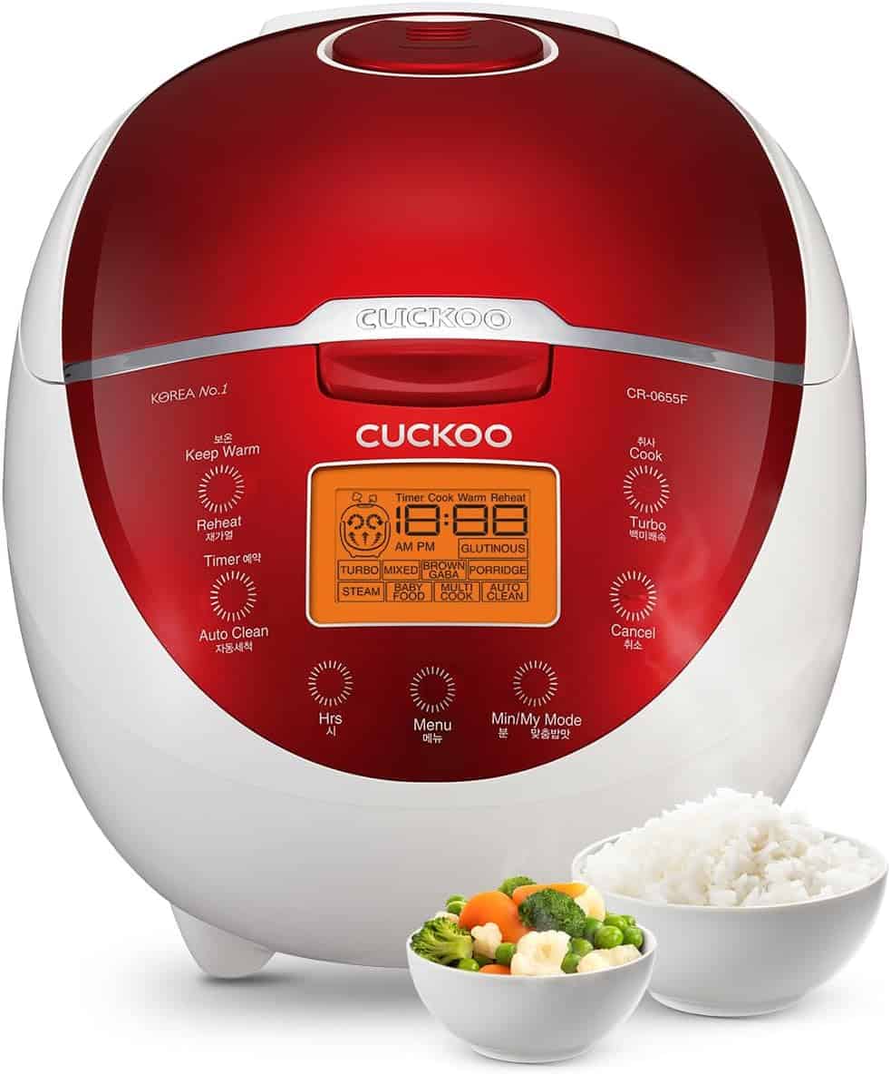 A red and white Cuckoo rice cooker with an orange digital display, shown with a bowl of cooked rice and a bowl of mixed vegetables in front.