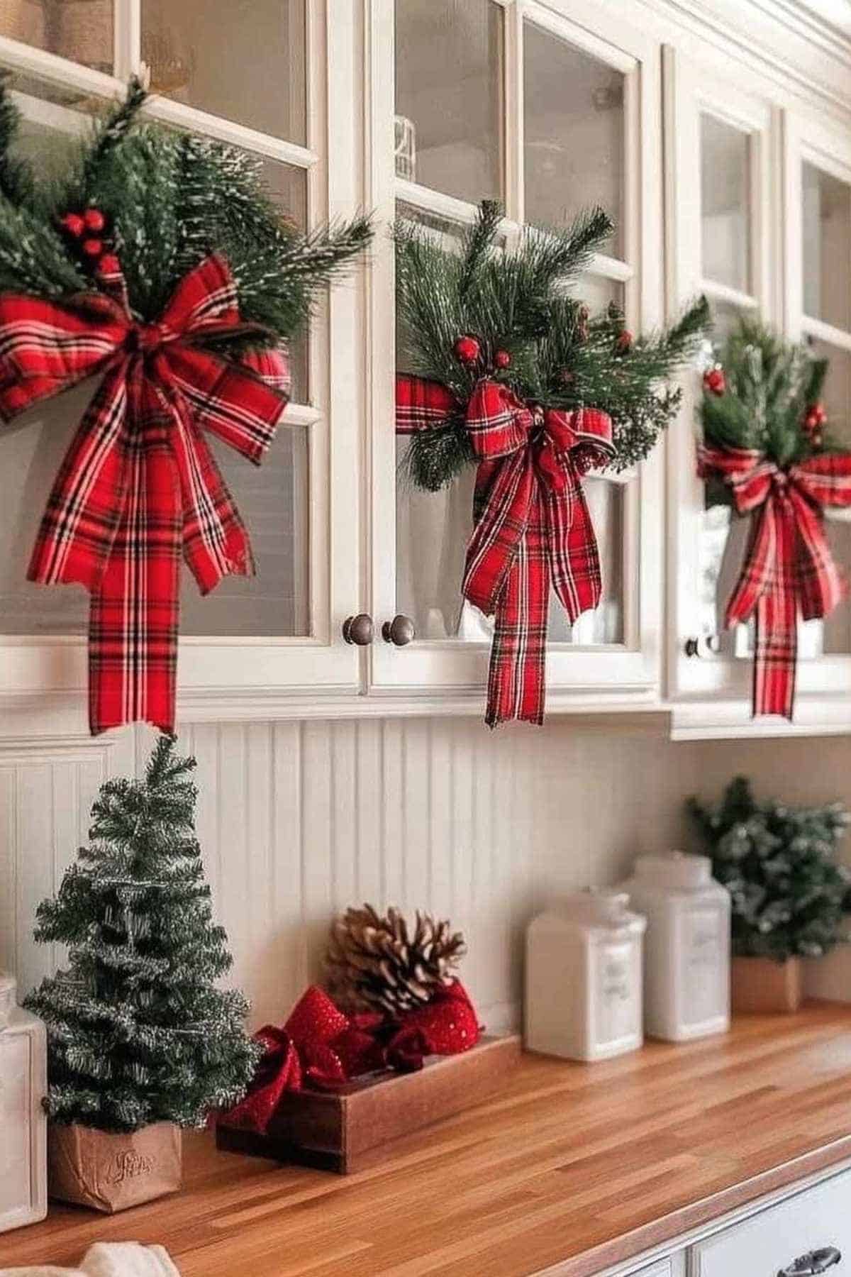 Three kitchen cabinet doors are decorated with greenery and large red plaid bows. A small artificial tree, pinecones, and canisters sit on the wooden countertop below.