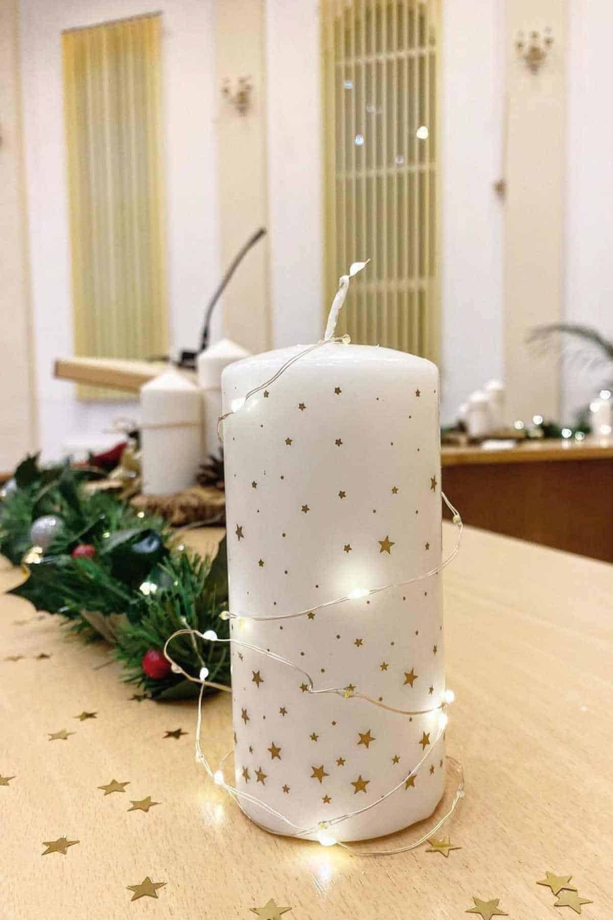 A white candle decorated with gold stars and wrapped in string lights sits on a wooden table with festive greenery in the background.