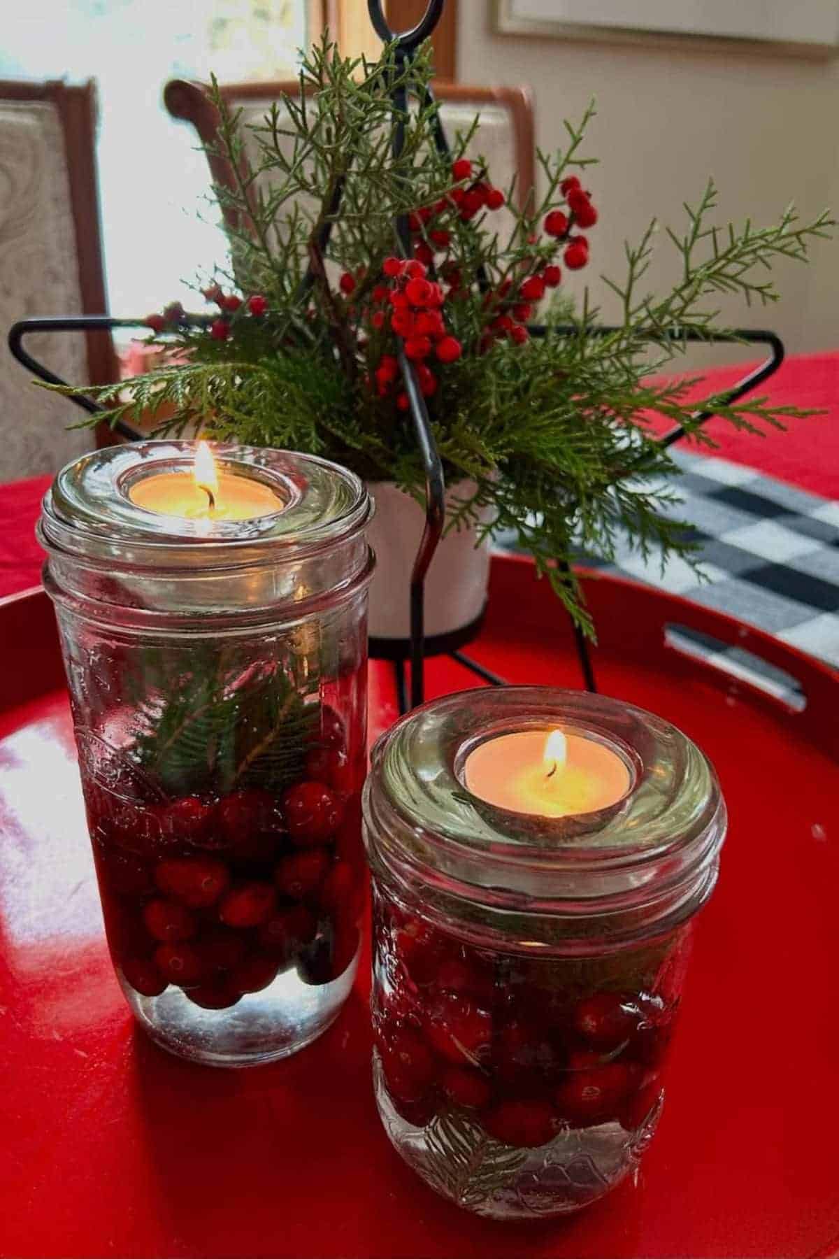 Two glass jars with floating candles, cranberries, and greenery sit on a red tray. A small plant with greenery and red berries is in the background.