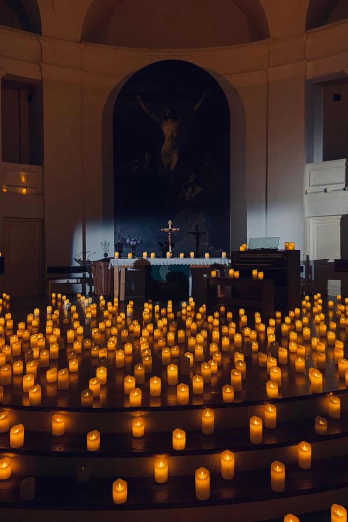A church altar is illuminated by numerous small candles arranged on steps, with a crucifix and religious painting in the background.