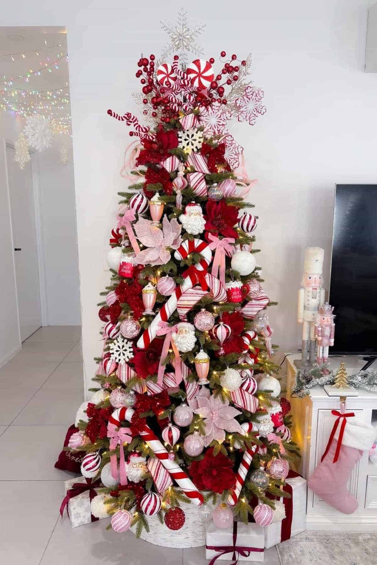 A decorated Christmas tree with red, white, and pink ornaments, candy cane decorations, and presents underneath, placed next to a white cabinet and a nutcracker figure.