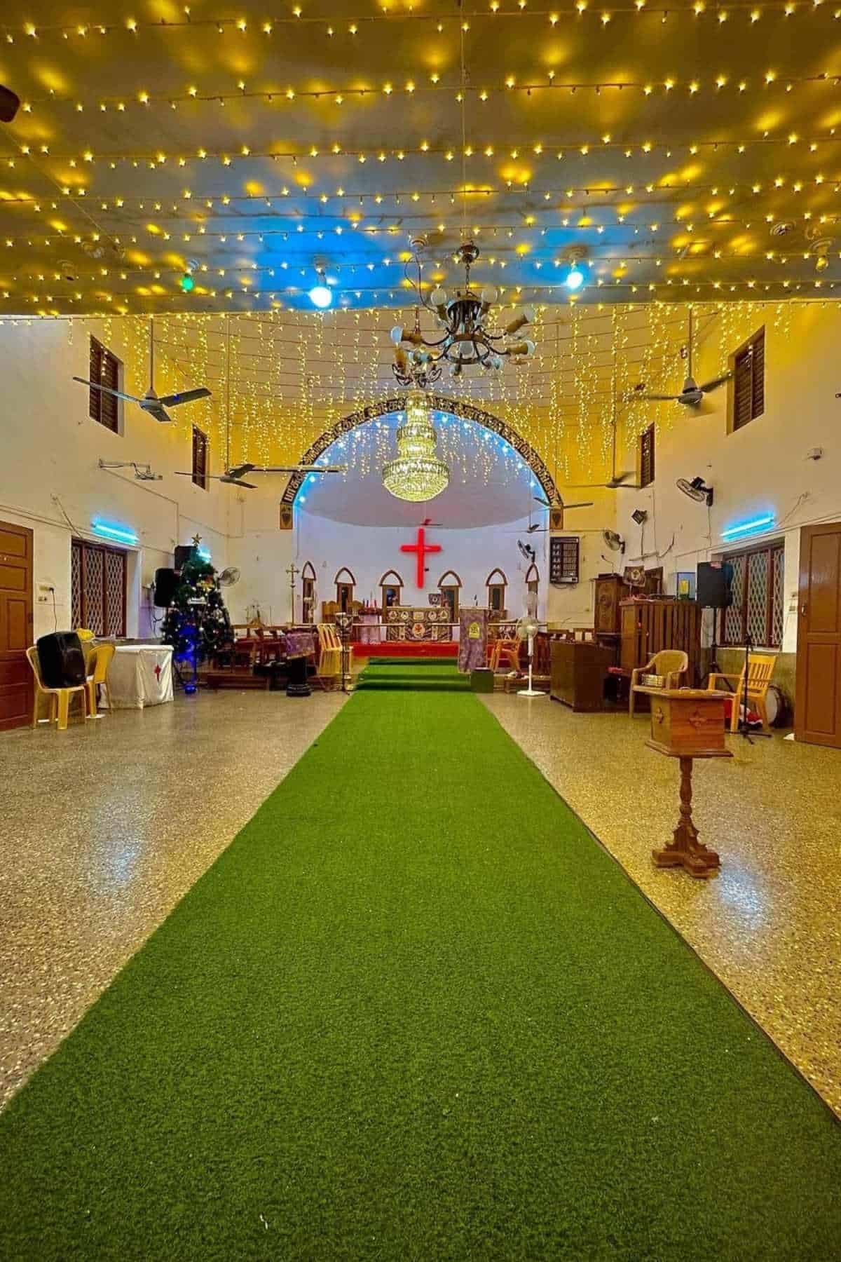 Interior of a church decorated with string lights, featuring a green carpet leading to the altar with a red cross, wooden chairs, and a chandelier overhead.