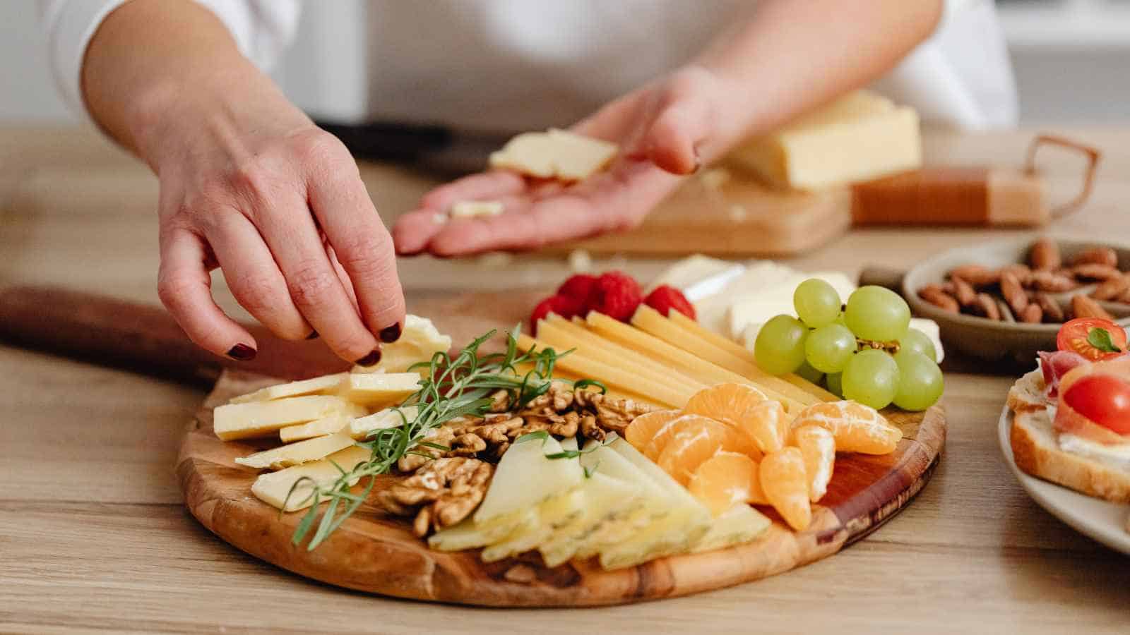 A person arranges cheese, grapes, oranges, walnuts, and herbs on a wooden serving board.
