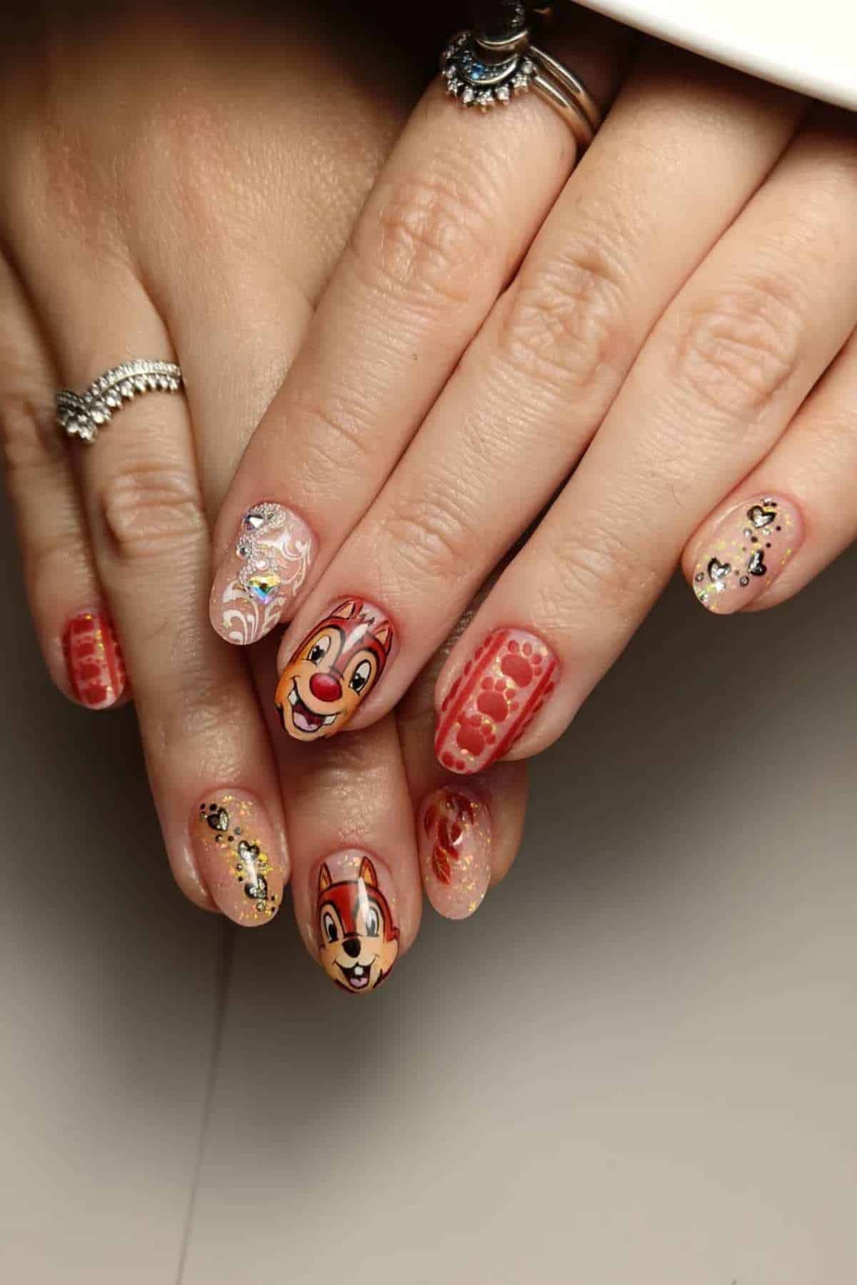 Close-up of hands with manicured nails featuring cartoon chipmunk faces, red patterns, glitter, and decorative designs; rings on fingers.