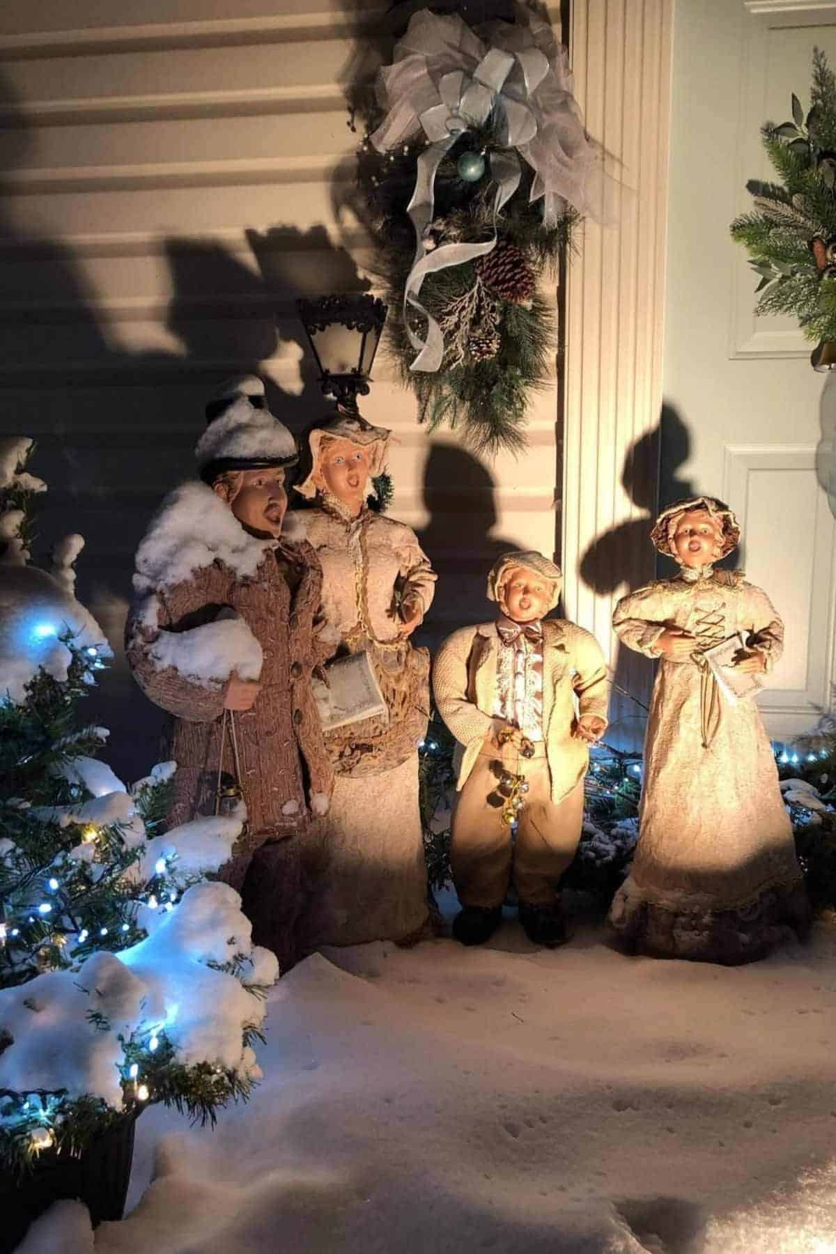 Four snow-covered caroler statues stand together by a decorated door, illuminated by holiday lights and surrounded by winter greenery and snow.