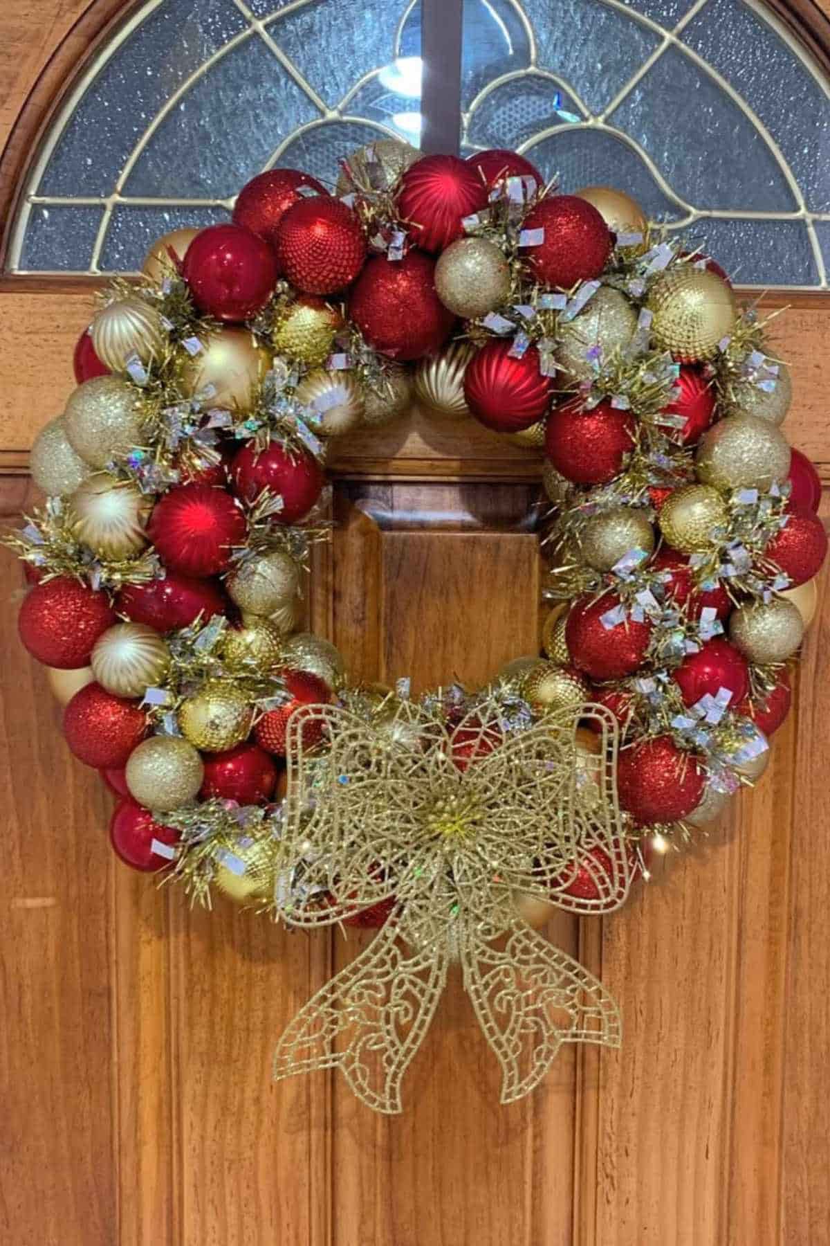 A wreath made of red and gold ornaments with small gold ribbons hangs on a wooden door, decorated with a large gold bow at the bottom.