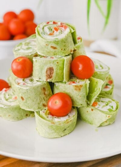 A plate of spinach tortilla Christmas pinwheels stacked in a pyramid shape, garnished with whole cherry tomatoes, on a wooden surface.