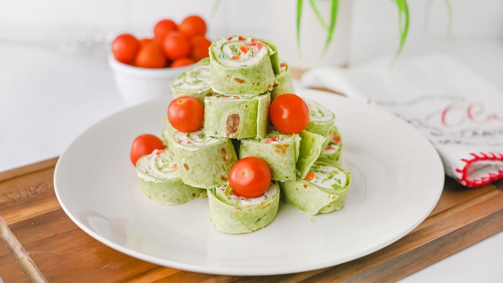 A plate of spinach tortilla Christmas pinwheels stacked in a pyramid shape, garnished with whole cherry tomatoes, on a wooden surface.