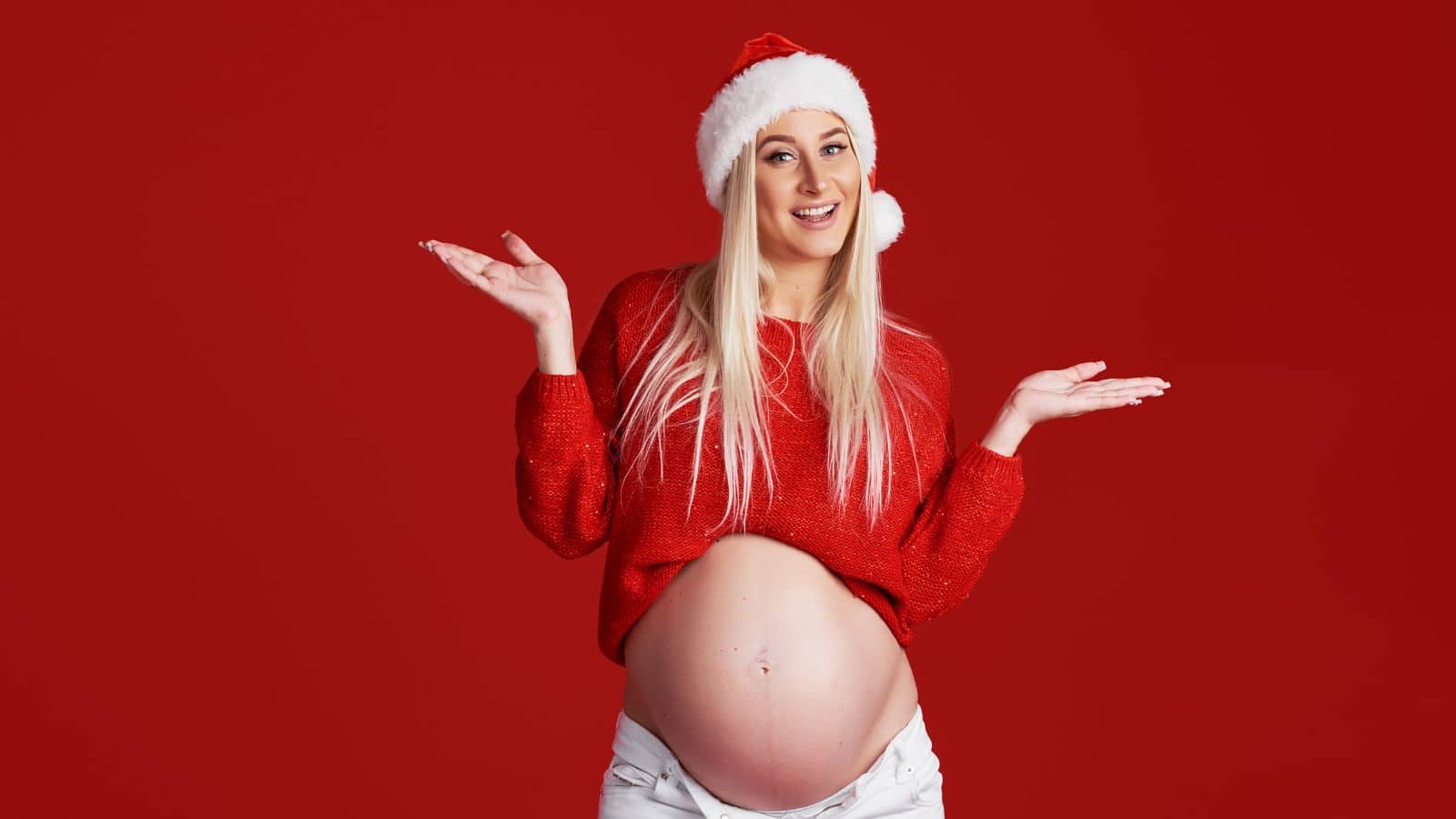 A pregnant woman in a red sweater and Santa hat smiles with her hands raised, standing in front of a solid red background.