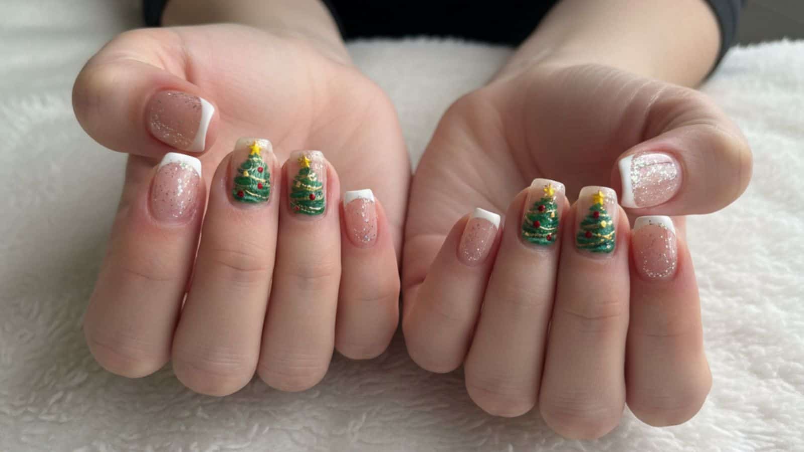 Hands with French tip nails, some decorated with painted Christmas trees and glitter, resting on a white surface.