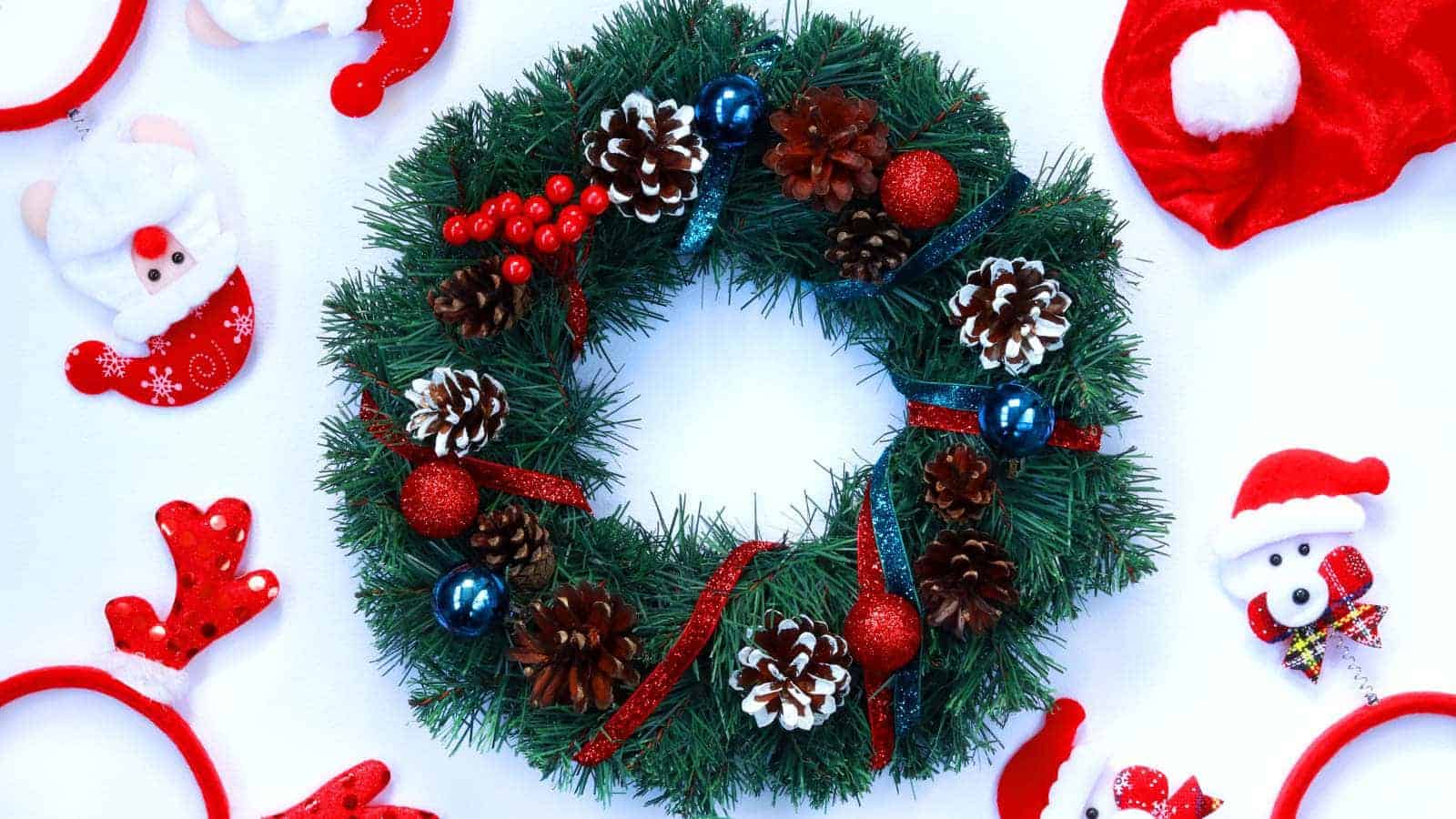 A Christmas wreath decorated with pinecones, red berries, and ornaments is surrounded by Santa-themed headbands and decorations on a white background.