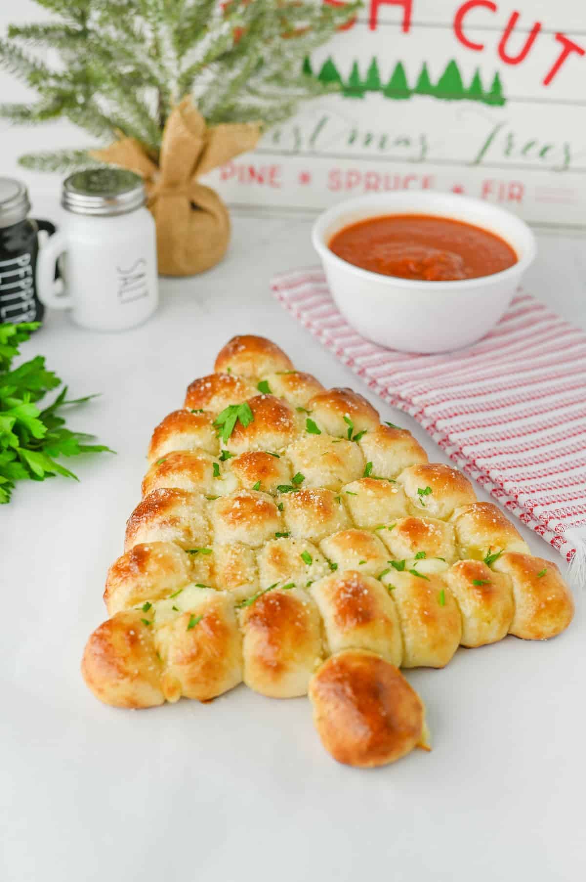 A Christmas tree-shaped pull-apart bread topped with parsley sits beside a bowl of marinara sauce, salt and pepper shakers, and a small potted pine.