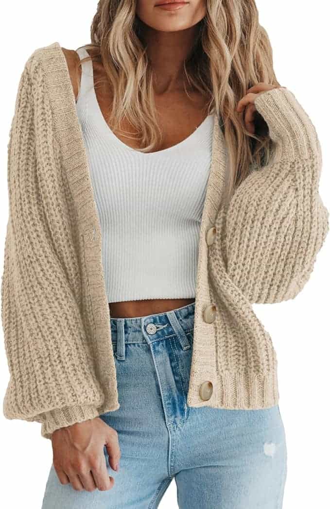 Woman wearing a beige chunky knit cardigan over a white ribbed crop top and high-waisted blue jeans, with loose wavy hair.