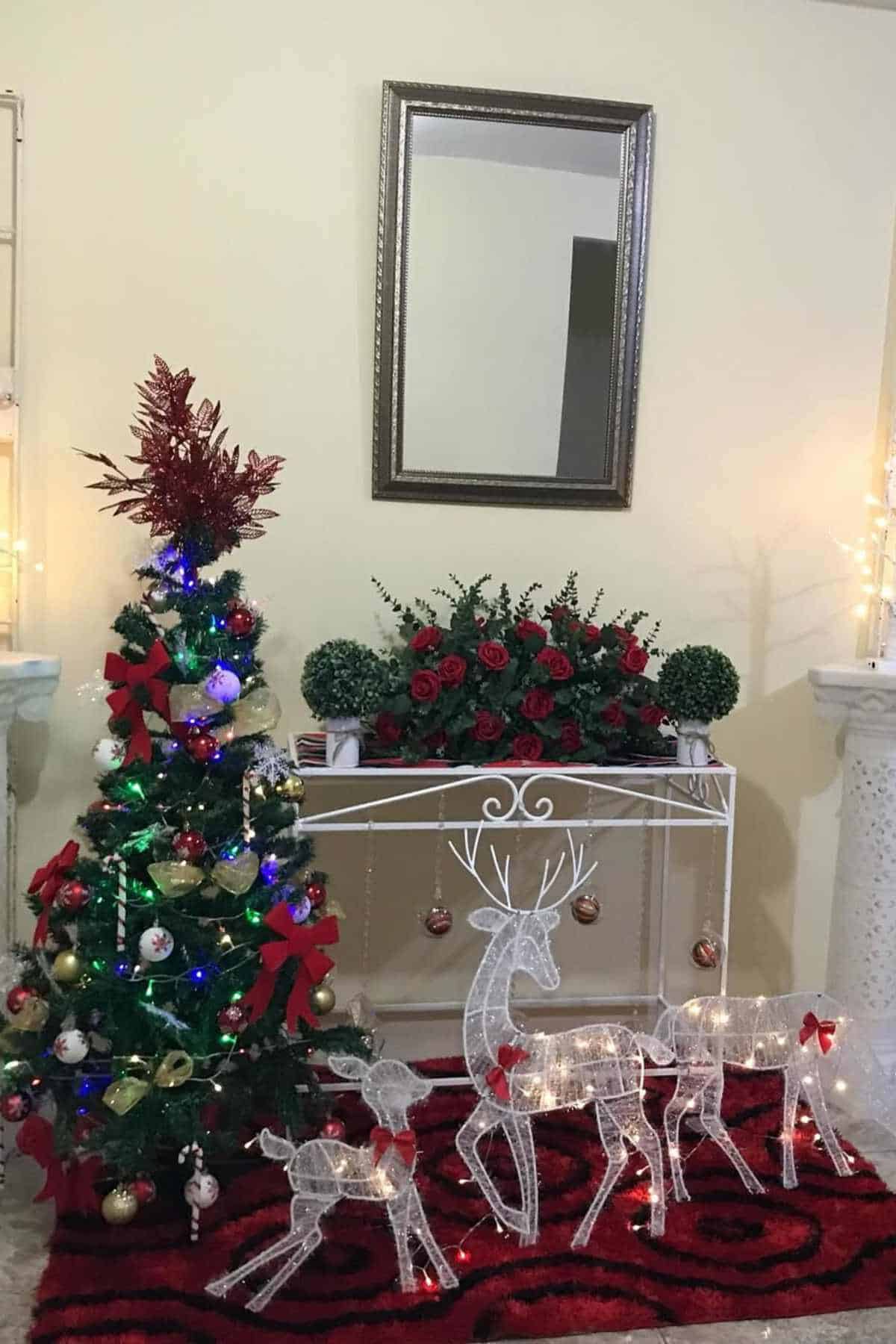 A decorated Christmas tree, three lighted reindeer figures, and a floral arrangement sit in front of a mirror on a red rug in a festive living room setting.