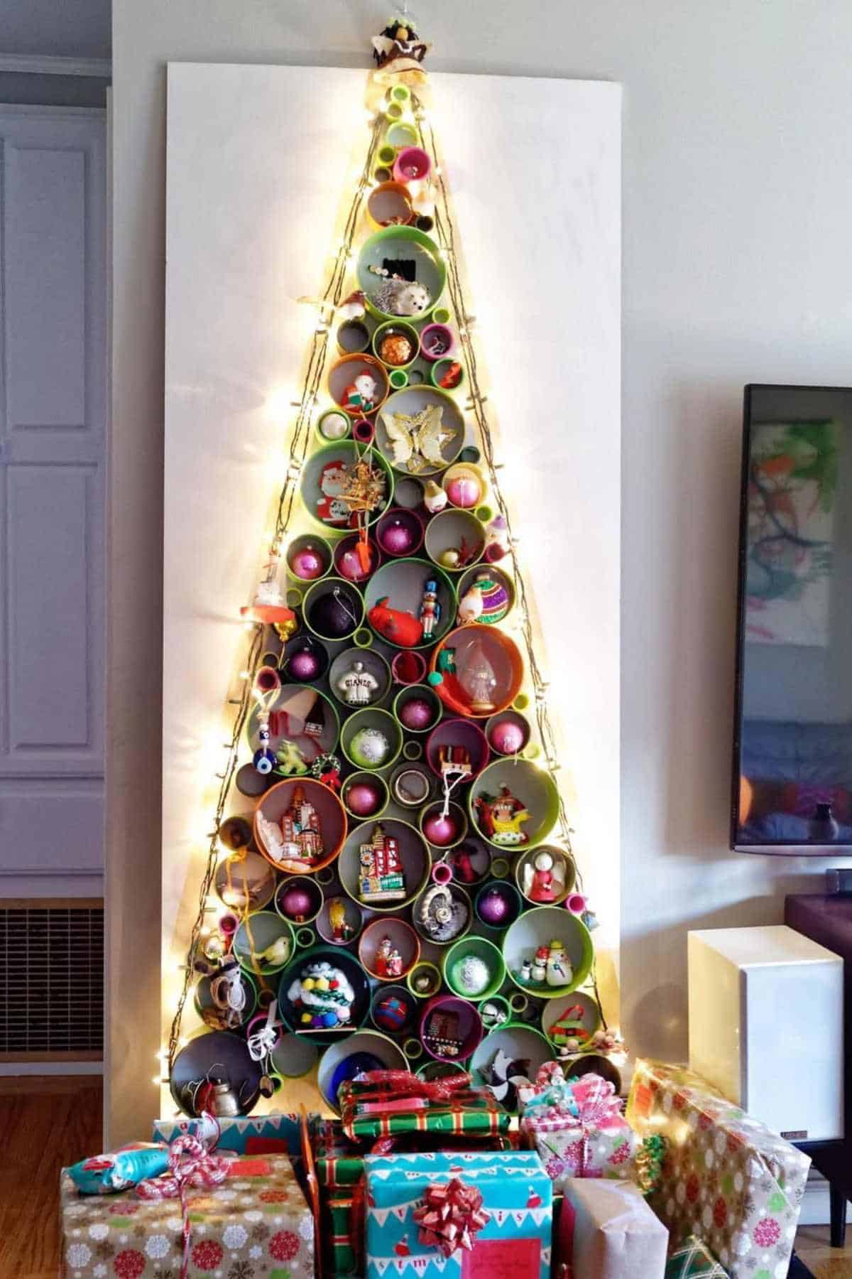 A wall-mounted Christmas tree made of circular shelves holds ornaments and is decorated with string lights, with wrapped presents arranged at the base.