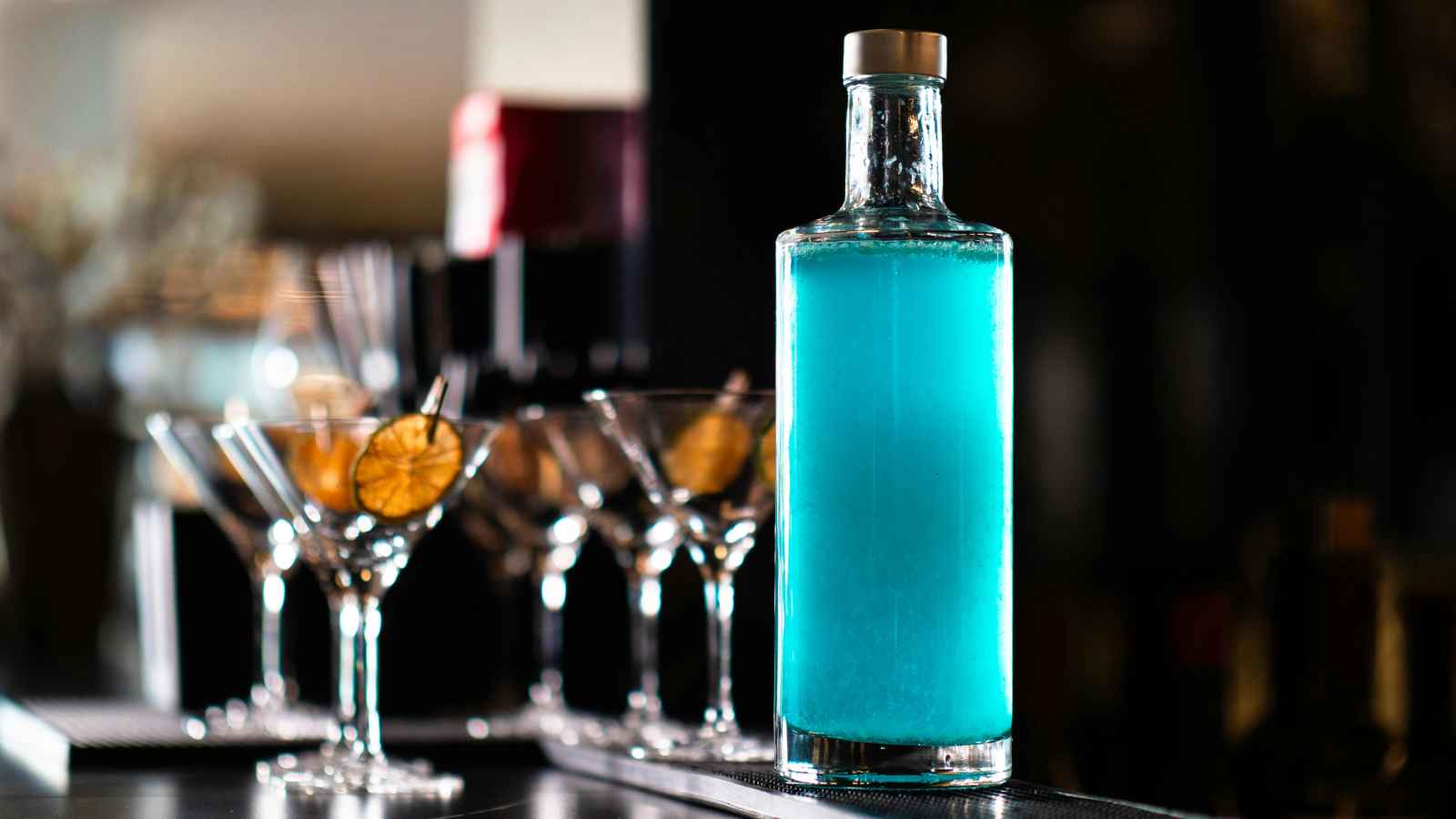 A tall glass bottle filled with bright blue liquid sits on a bar counter next to several martini glasses garnished with dried lime slices.