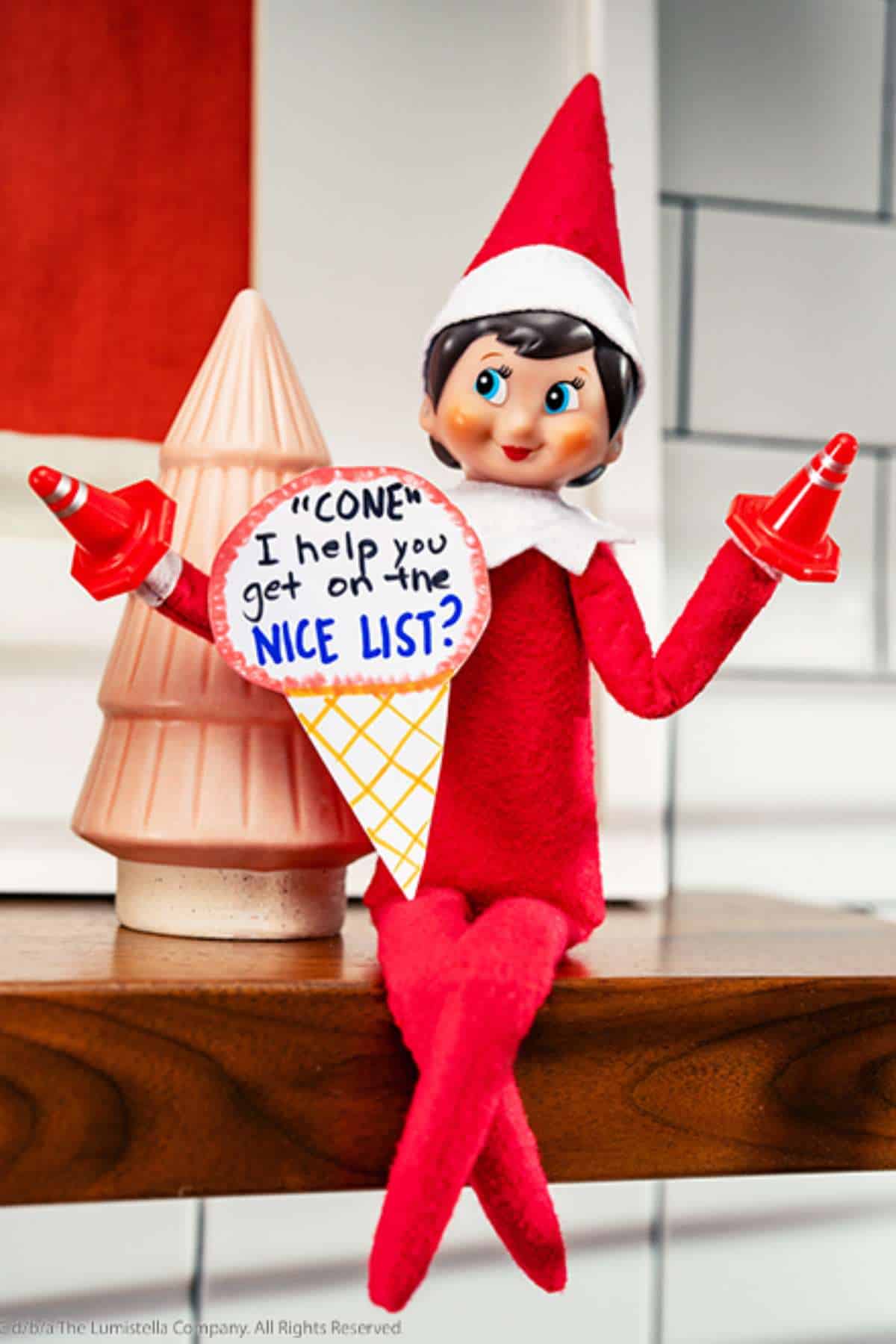 Elf on the Shelf doll sitting on a shelf, holding an ice cream cone-shaped sign that says "CONE I help you get on the NICE LIST?" and two red traffic cone on its fingers.