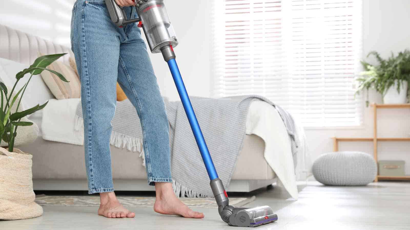 Person using a cordless stick vacuum to clean the floor in a bright, modern bedroom with a bed, basket plant, and natural light from the window.