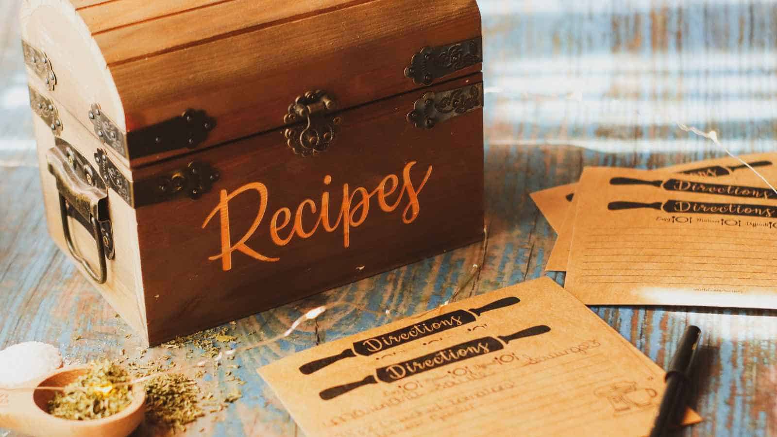 A wooden recipe box labeled "Recipes" sits on a table next to recipe cards, a pen, and scattered herbs.