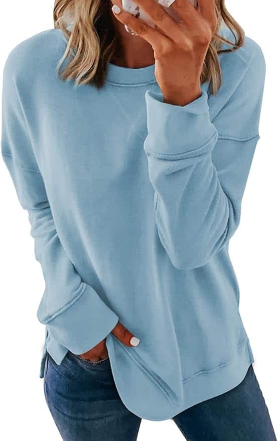 A person wearing a light blue long-sleeve sweatshirt and blue jeans stands with one hand in a pocket and the other hand held near their face.