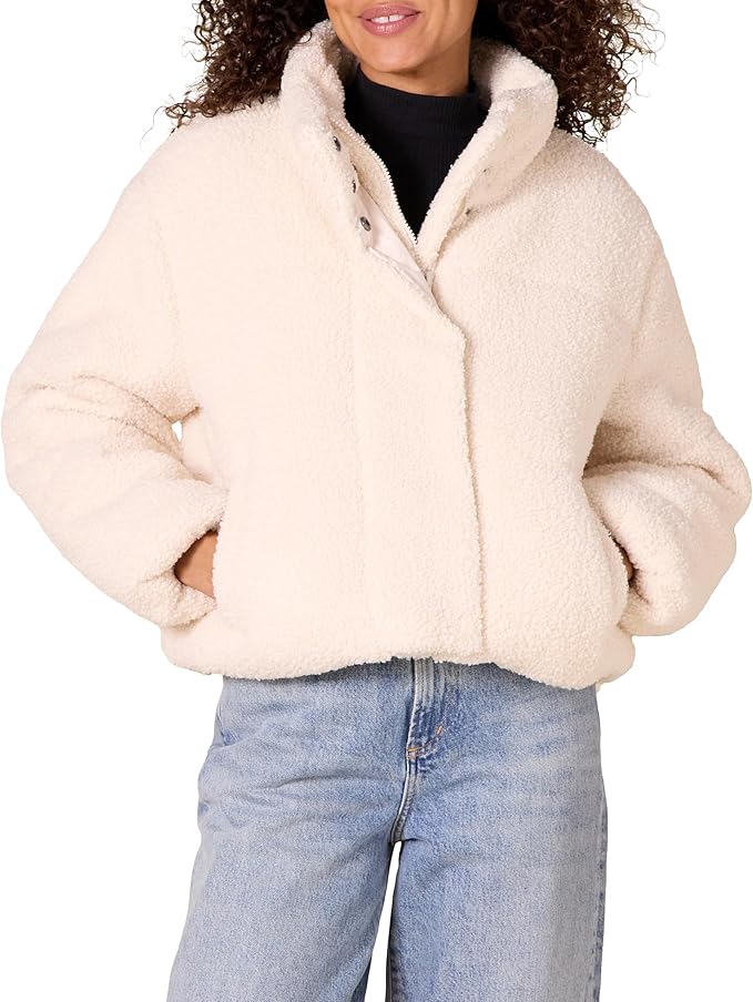 A person wearing a cream-colored, fuzzy sherpa jacket over a black top and light blue jeans, with hands in the jacket pockets.