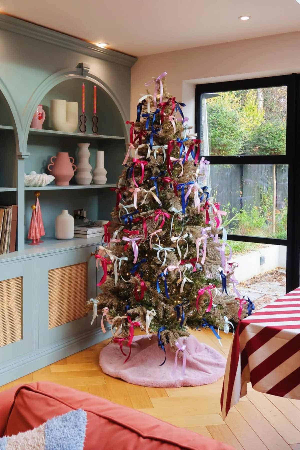 A Christmas tree stands in a modern living room, decorated with numerous colorful bows and topped with a pink cloth base. Shelves with vases and books are in the background.
