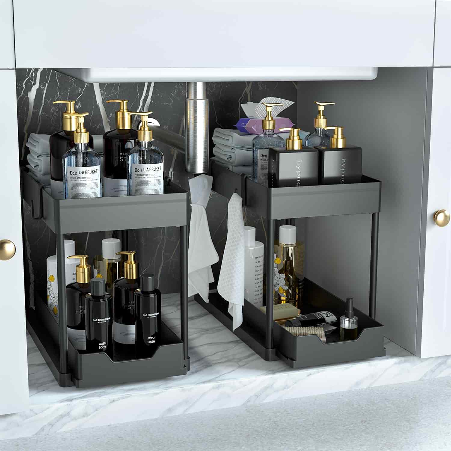 Two black tiered organizers under a sink hold bottles, rolled towels, and cleaning supplies on a marble-patterned surface with a visible pipe in the background.