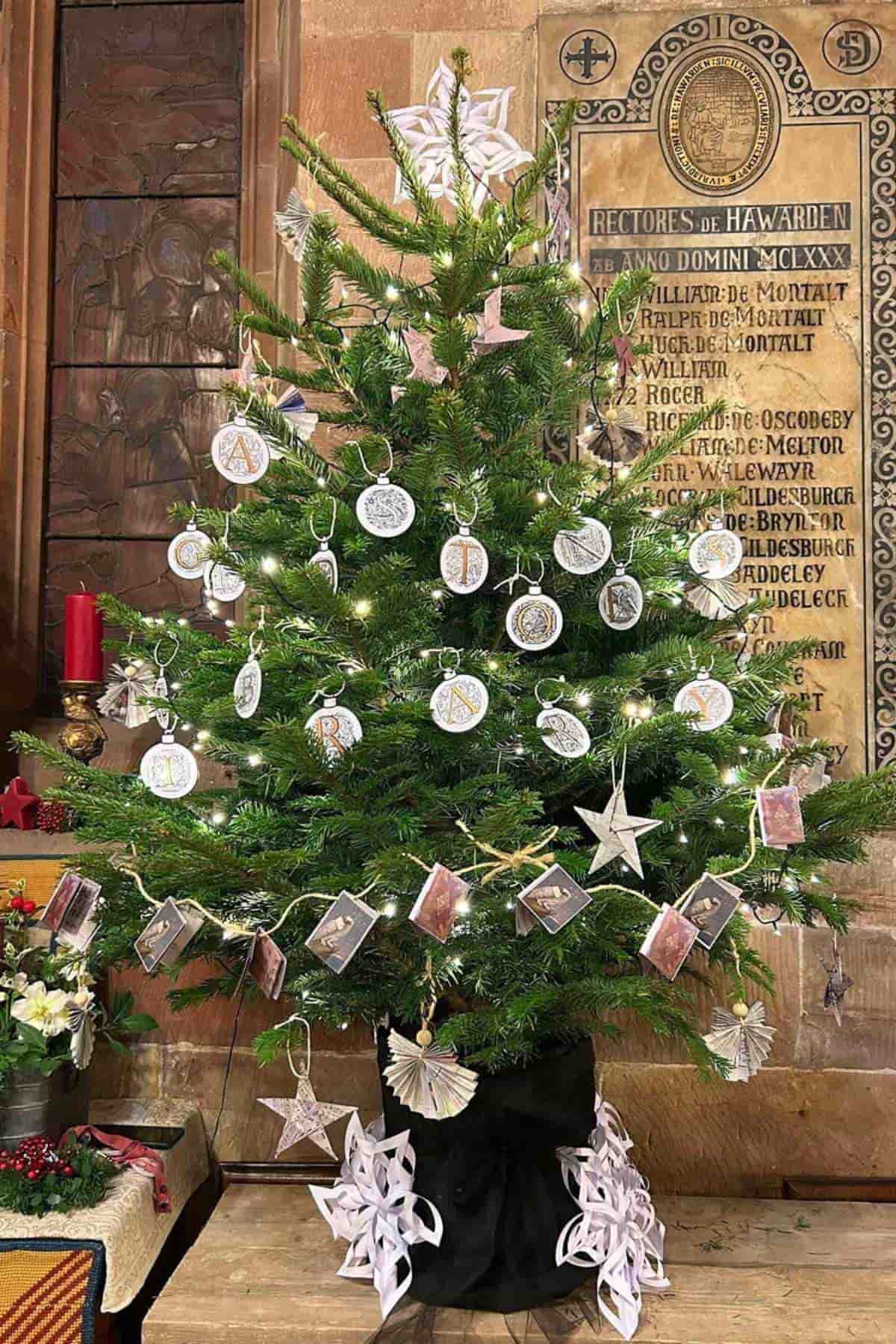 Decorated Christmas tree with white and silver ornaments, paper stars, and photo cards, placed indoors against a stone wall with engraved plaques.