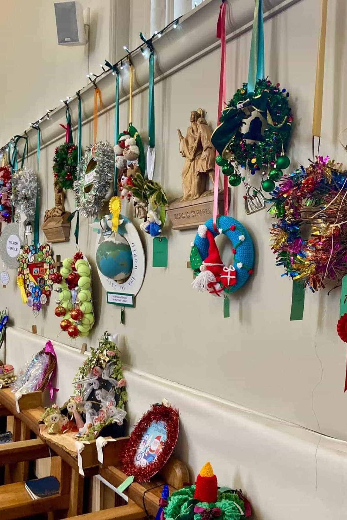A variety of handmade holiday wreaths and decorations hang on a wall above wooden chairs in a room.