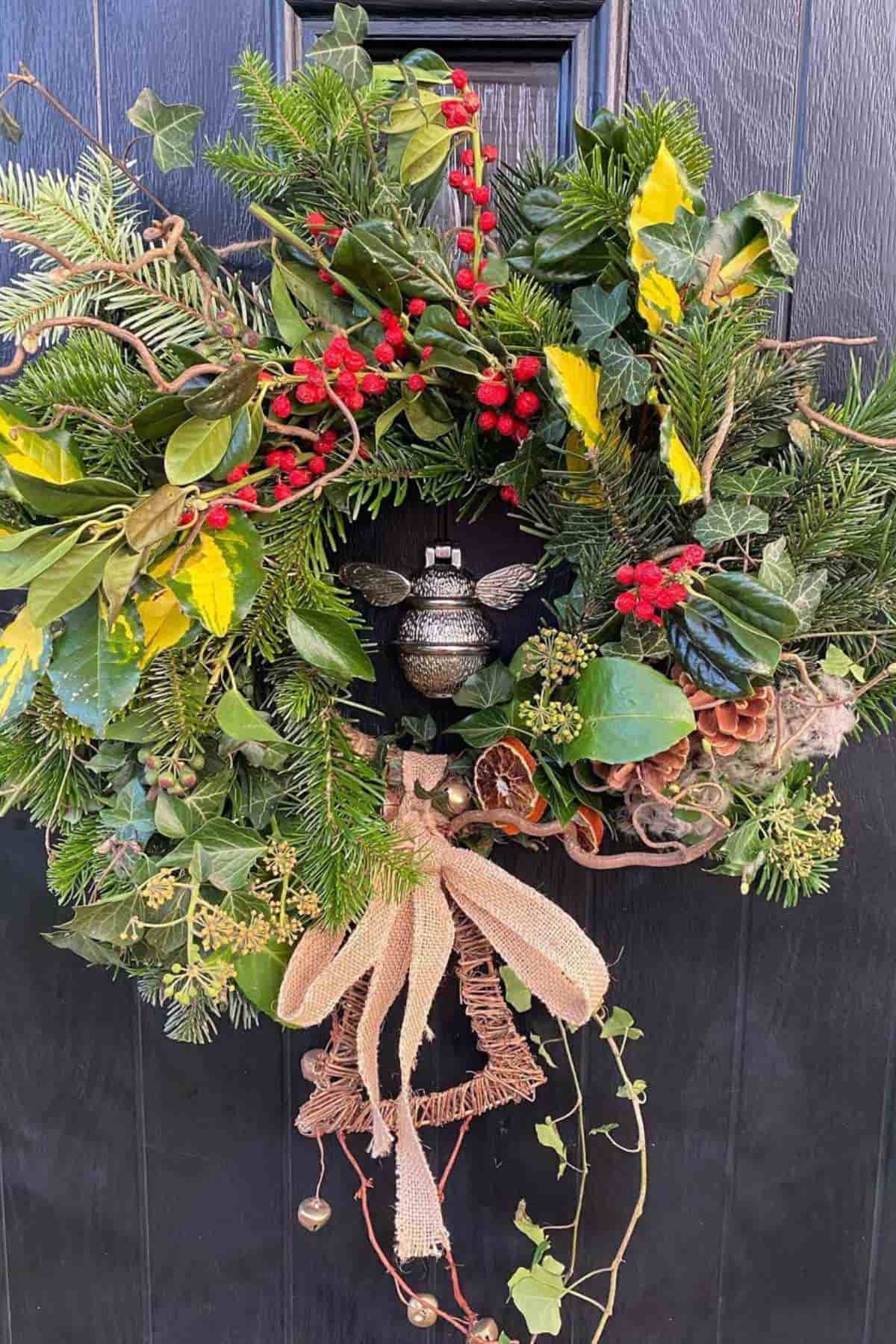 A festive wreath made of pine, holly, red berries, and greenery with a burlap bow hangs on a dark door.