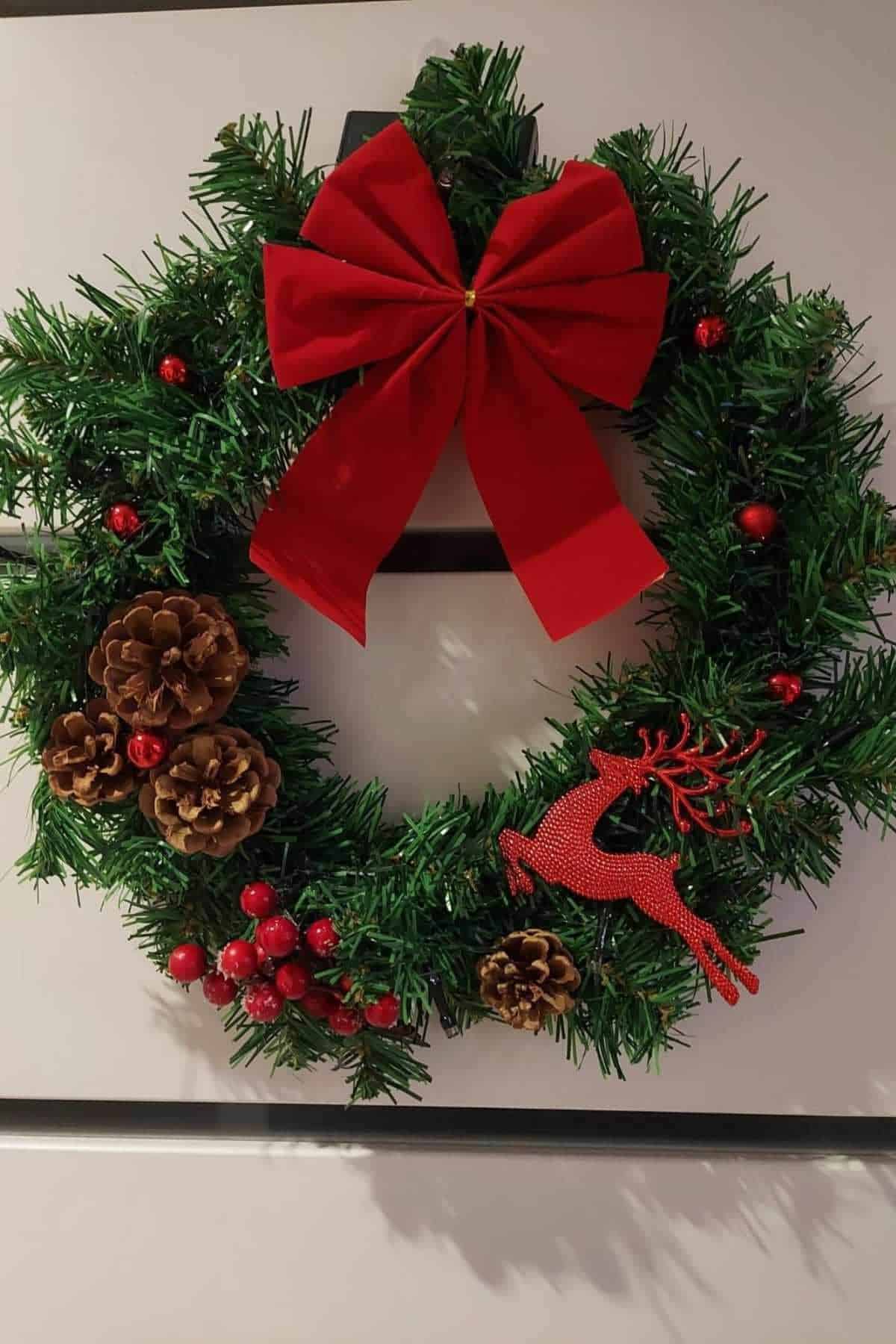 A Christmas wreath decorated with a large red bow, pinecones, red berries, and a red reindeer ornament, hanging on a white door.