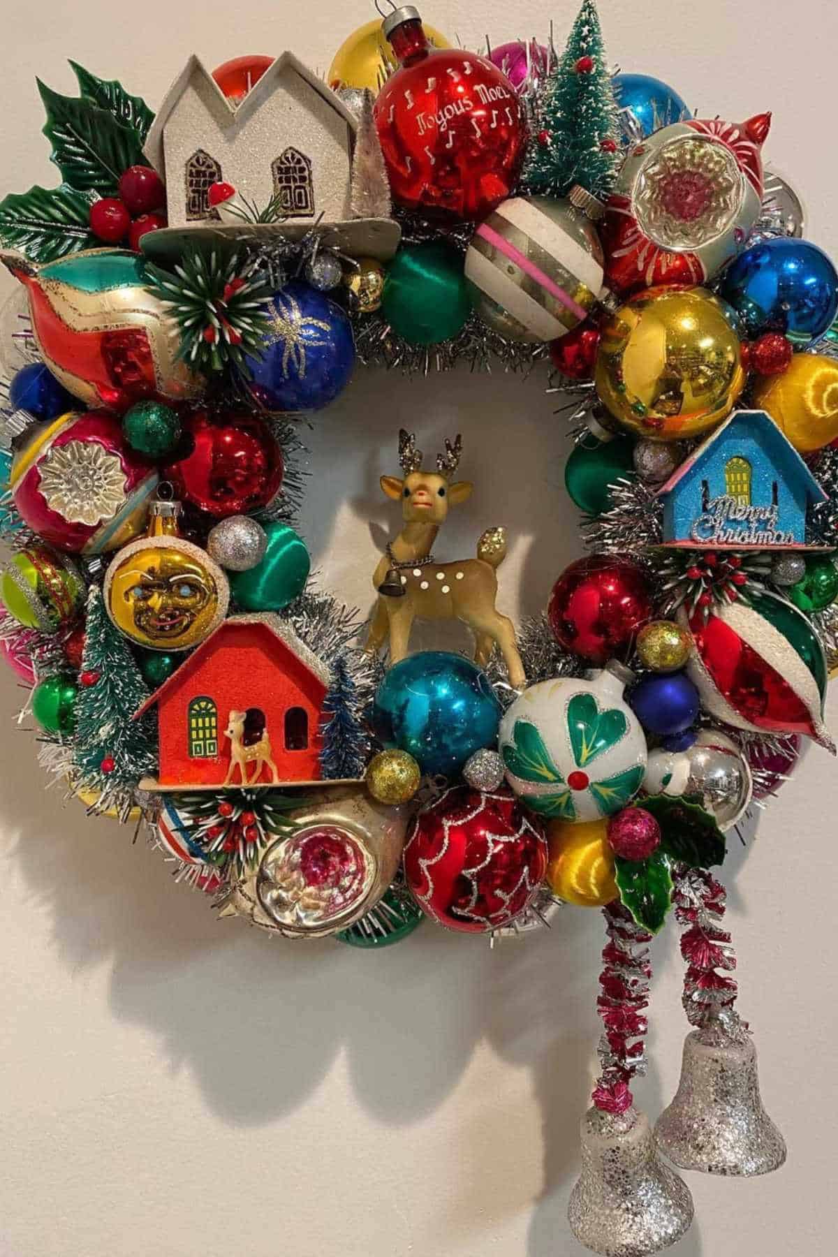 A colorful Christmas wreath decorated with vintage ornaments, small houses, baubles, a reindeer figure, tinsel, and two hanging silver bells.