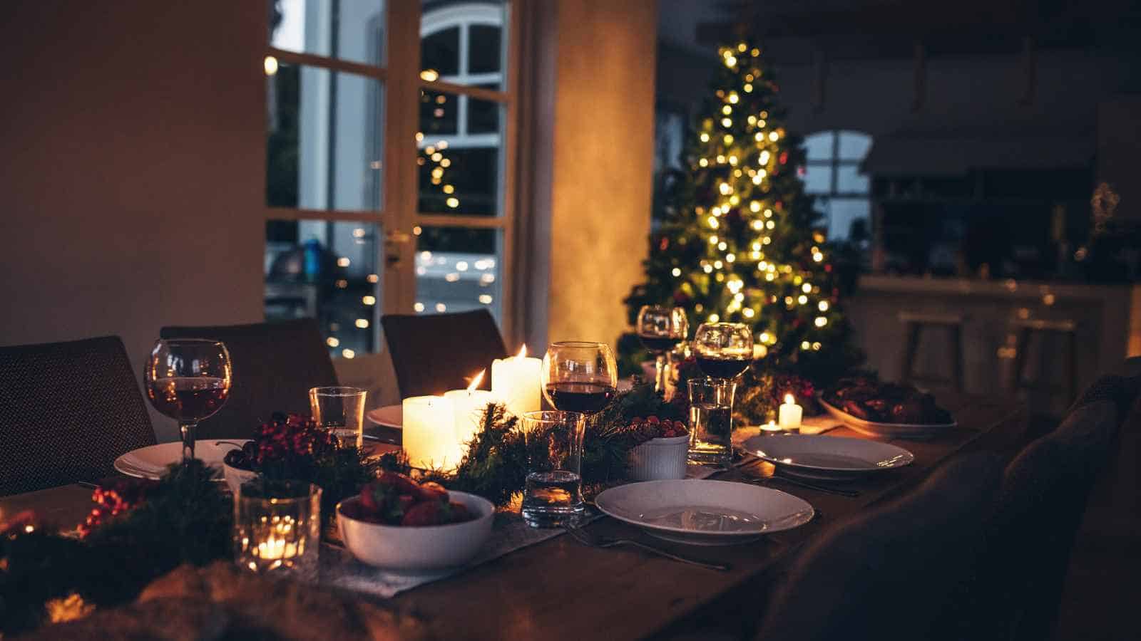 A dining table set for a meal with candles, plates, glasses, and food, with a decorated Christmas tree and string lights in the background.
