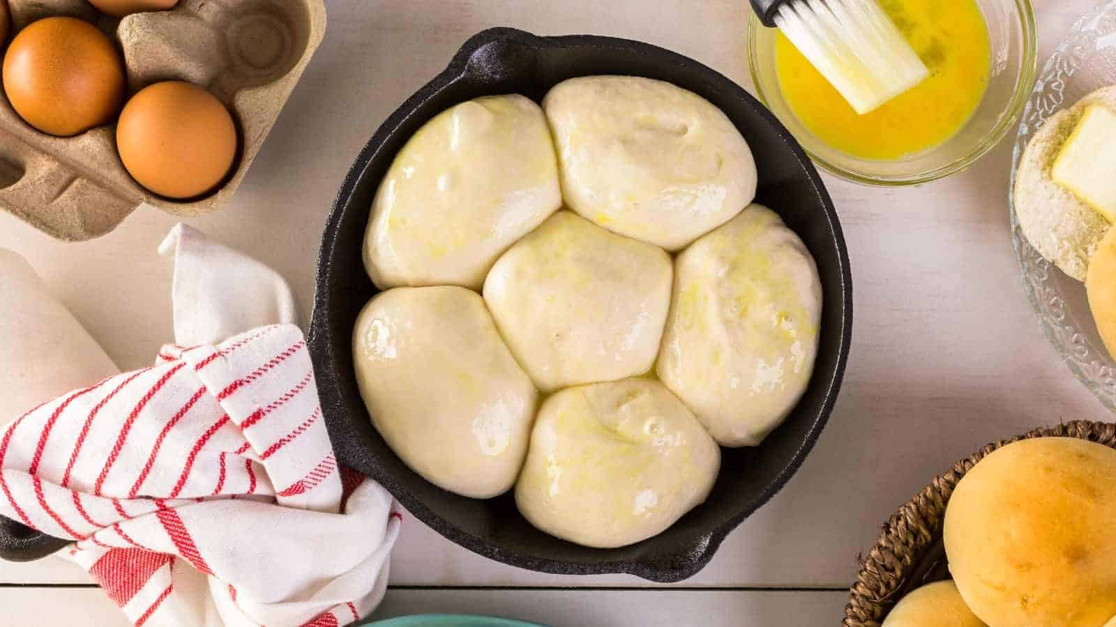 A cast iron skillet filled with seven unbaked dough balls brushed with egg wash, surrounded by eggs, melted butter, and a kitchen towel.